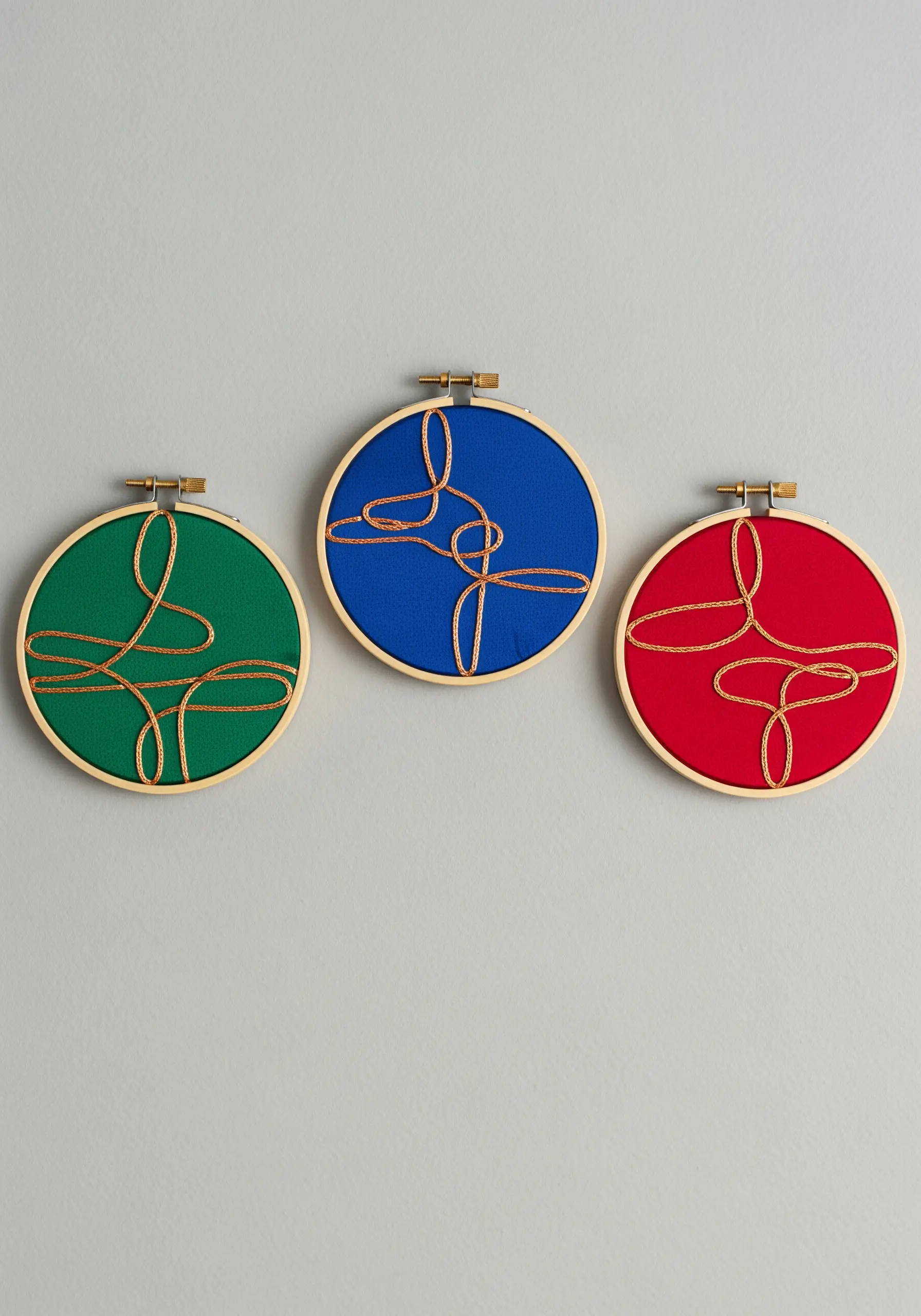 Three embroidery hoops in red, green, and blue, each with a single gold cord couched on top.