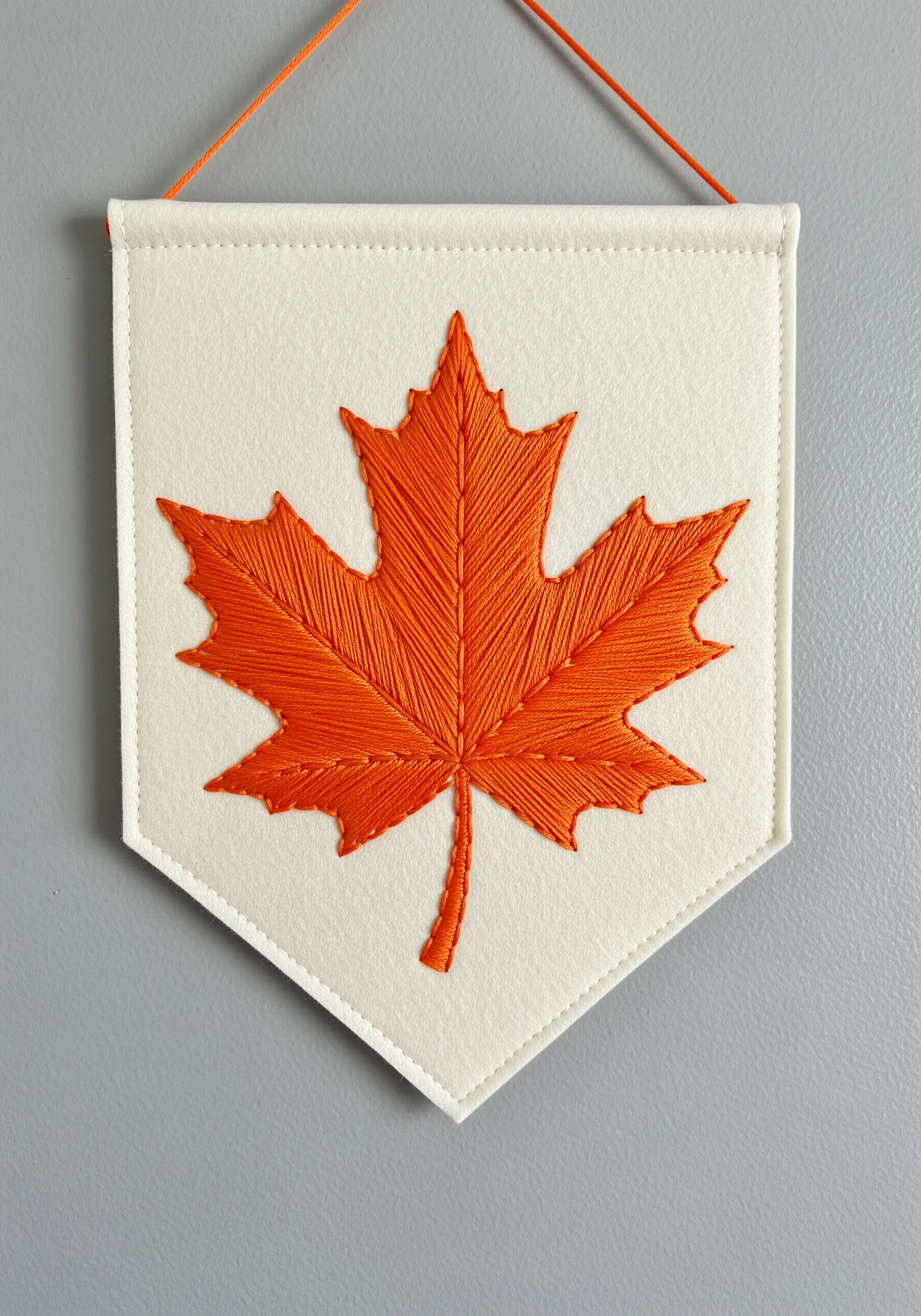 A bright orange maple leaf with directional satin stitch on a white felt banner.