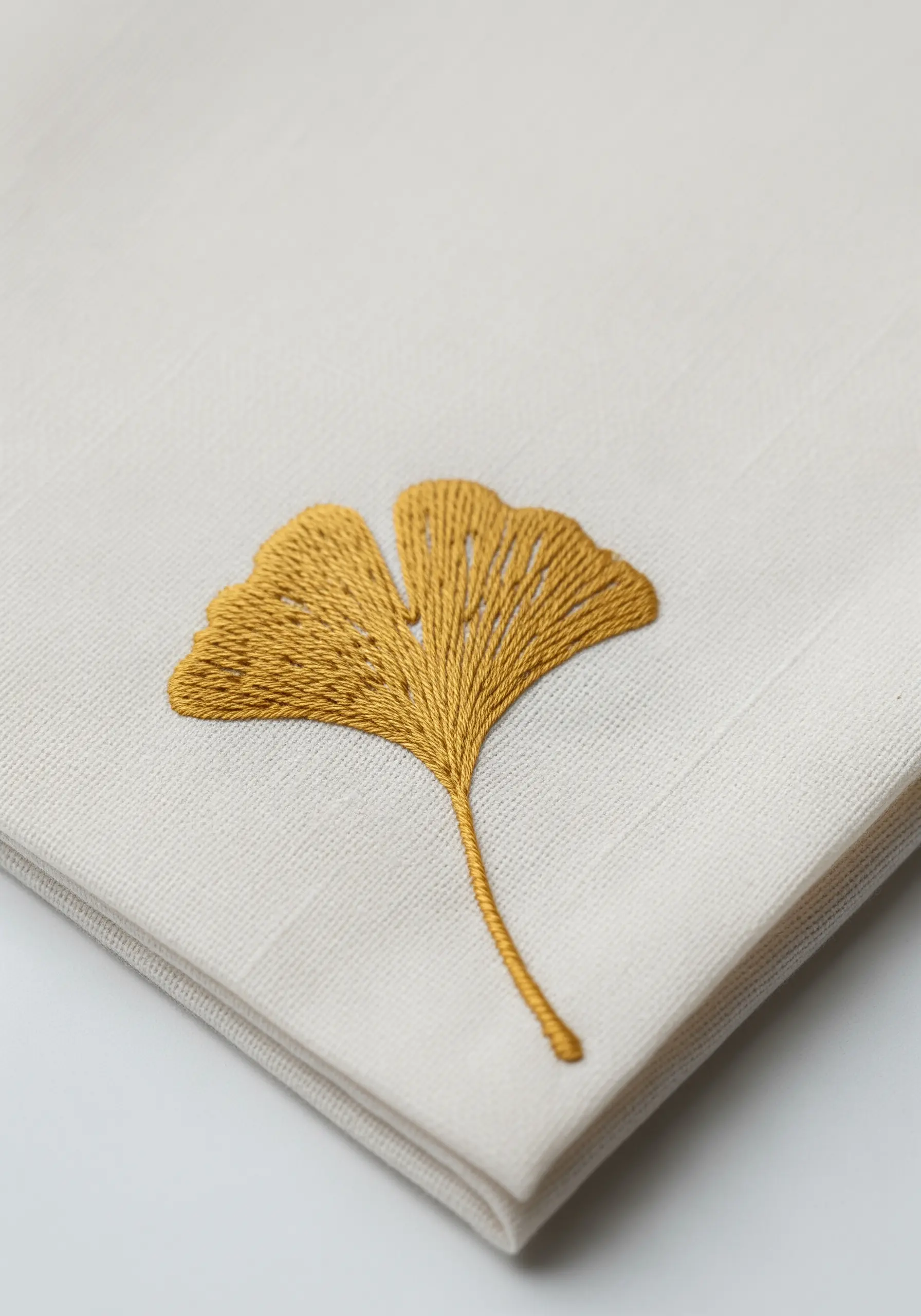 A golden-yellow ginkgo leaf embroidered with directional satin stitch on a cream handkerchief.