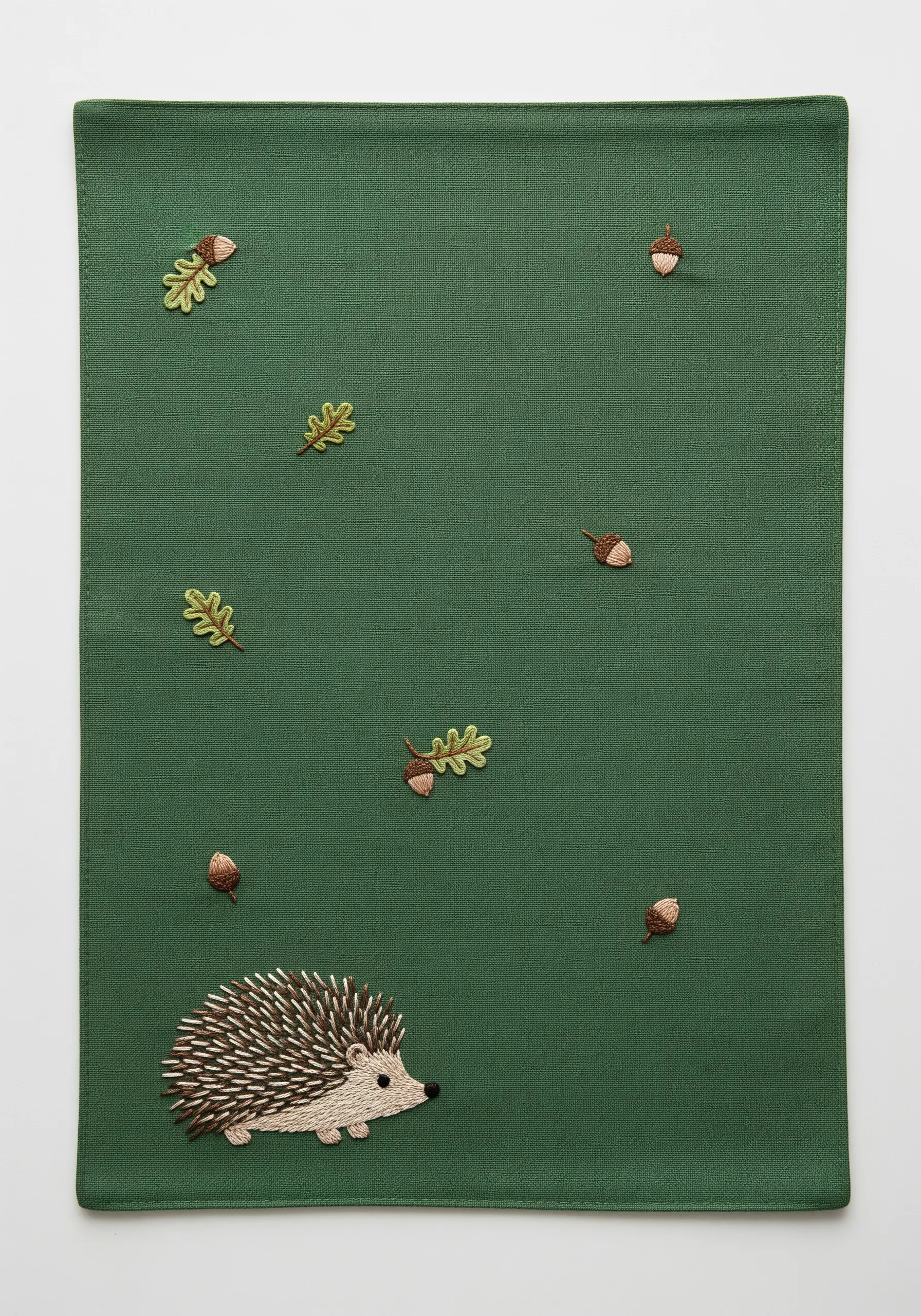A small hedgehog embroidered in the corner of a green placemat with scattered acorns and leaves.