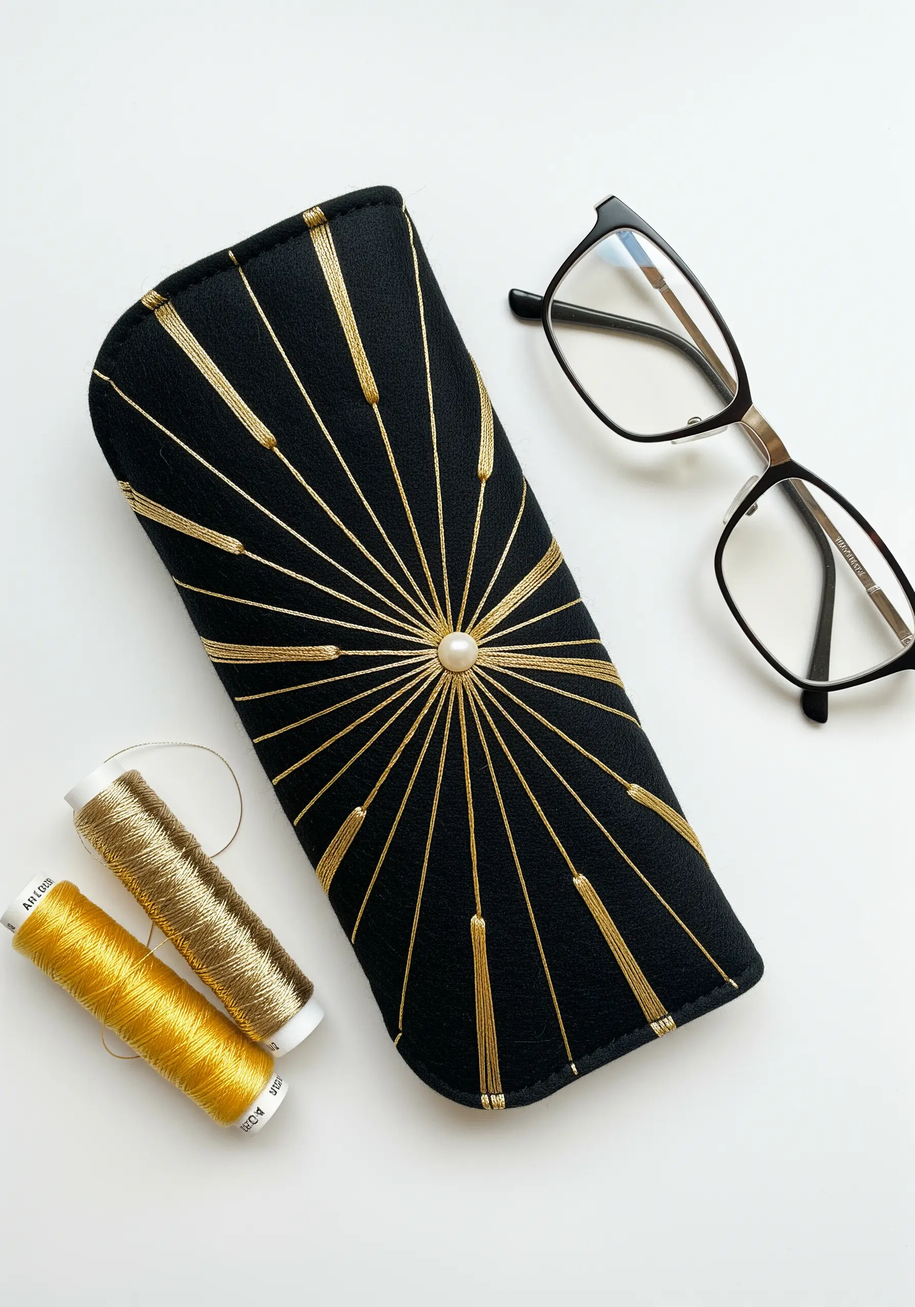 A black glasses case with a gold sunburst embroidery pattern radiating from a central pearl.