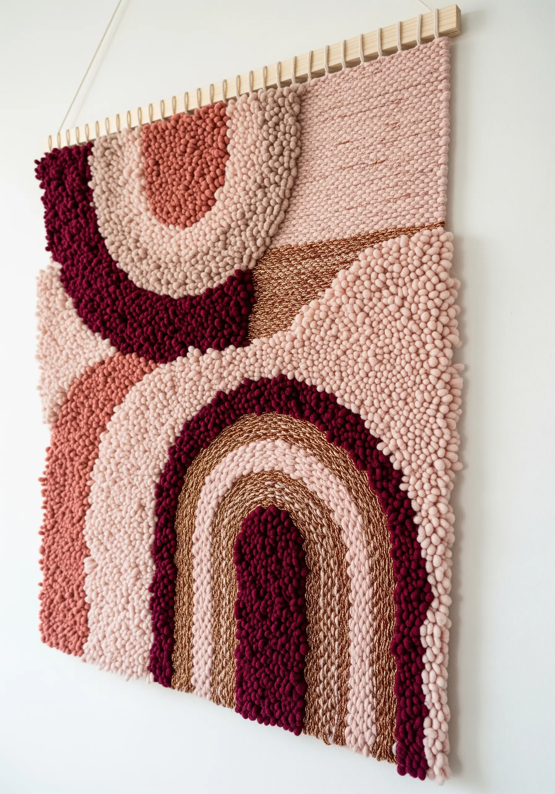 A textured wall hanging with punch needle loops and woven arches in pinks and reds.