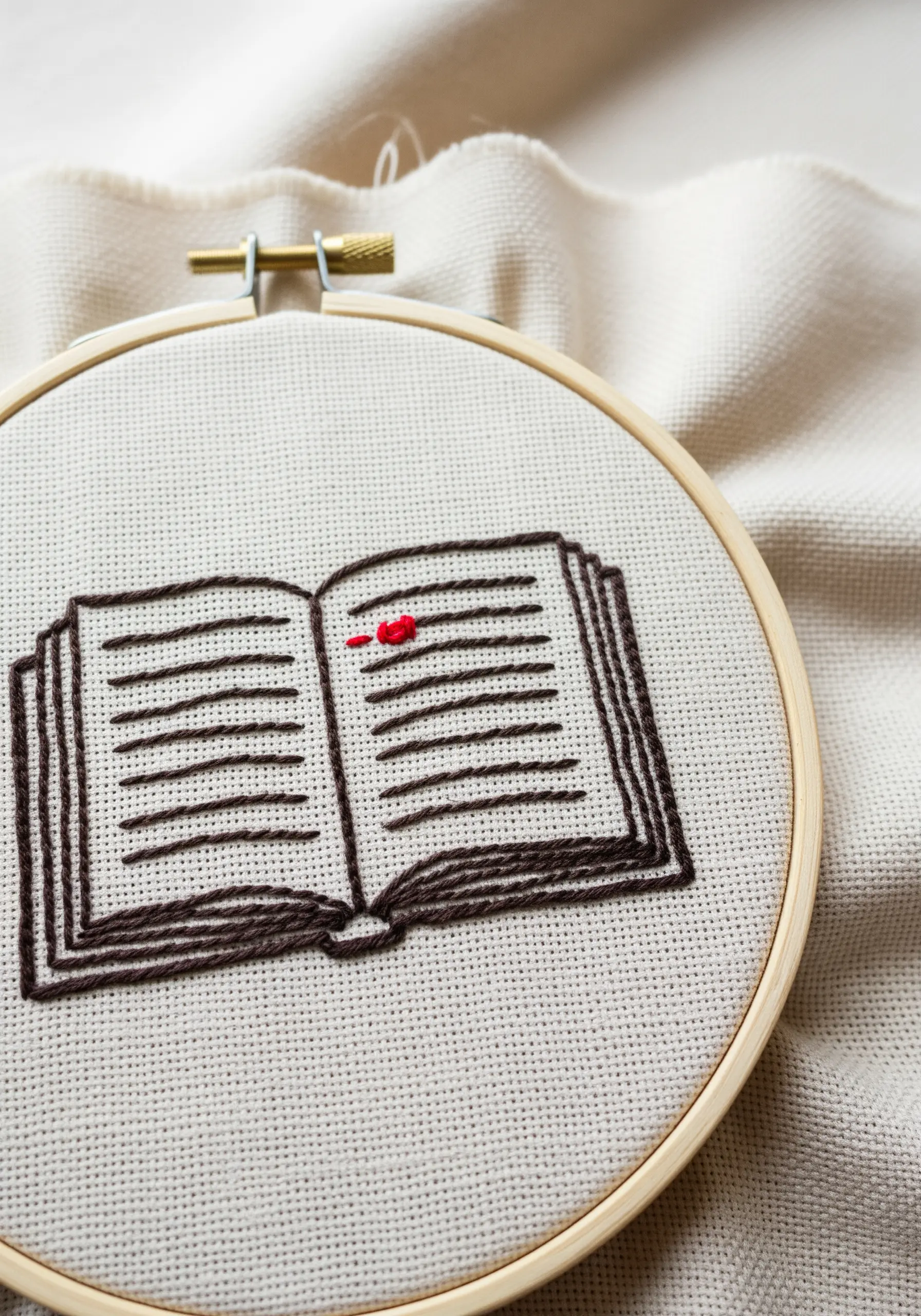 Embroidered open book with a single red French knot highlighting a line of text.