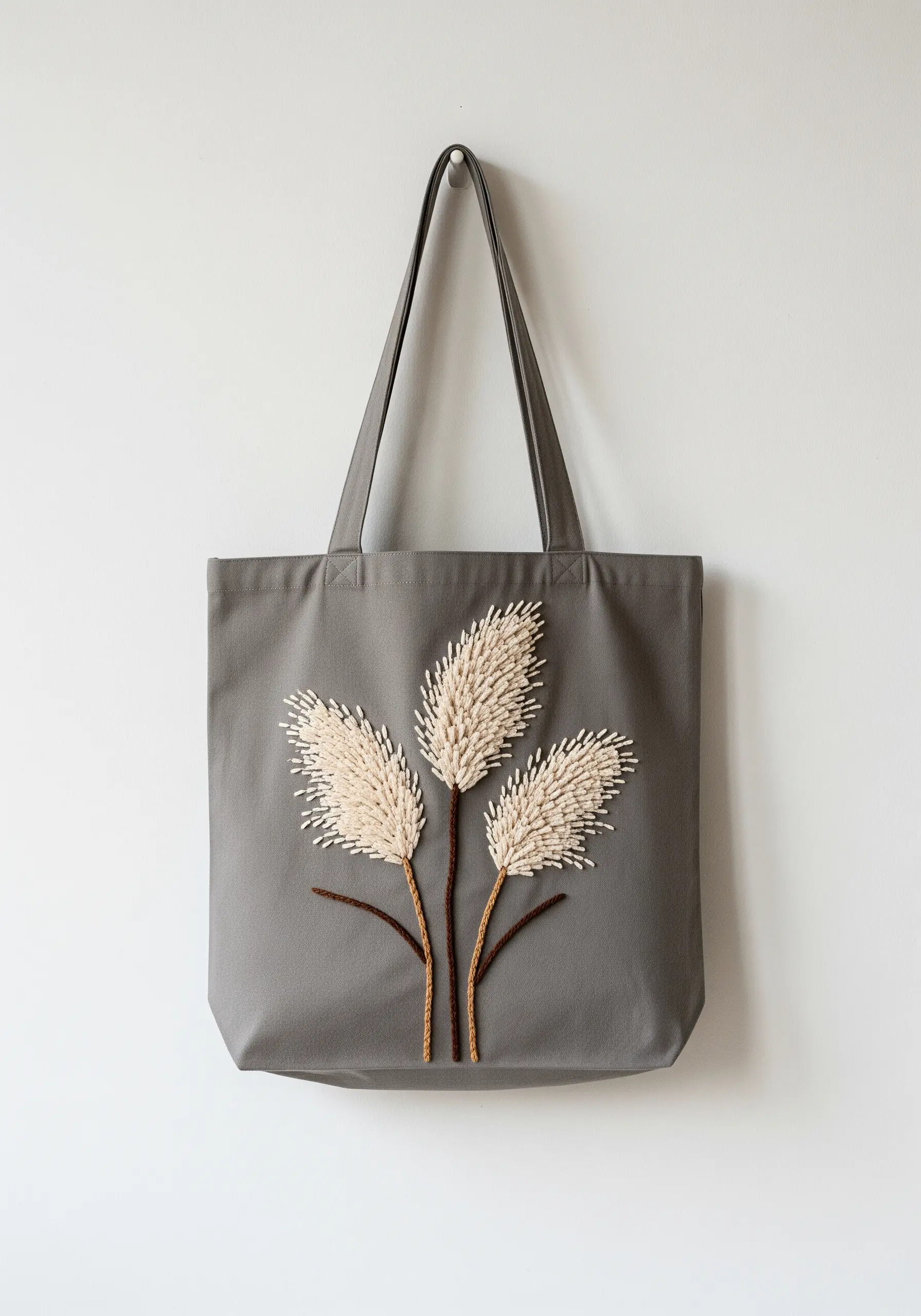 Three stalks of pampas grass with fluffy, textured embroidery on a gray tote bag.