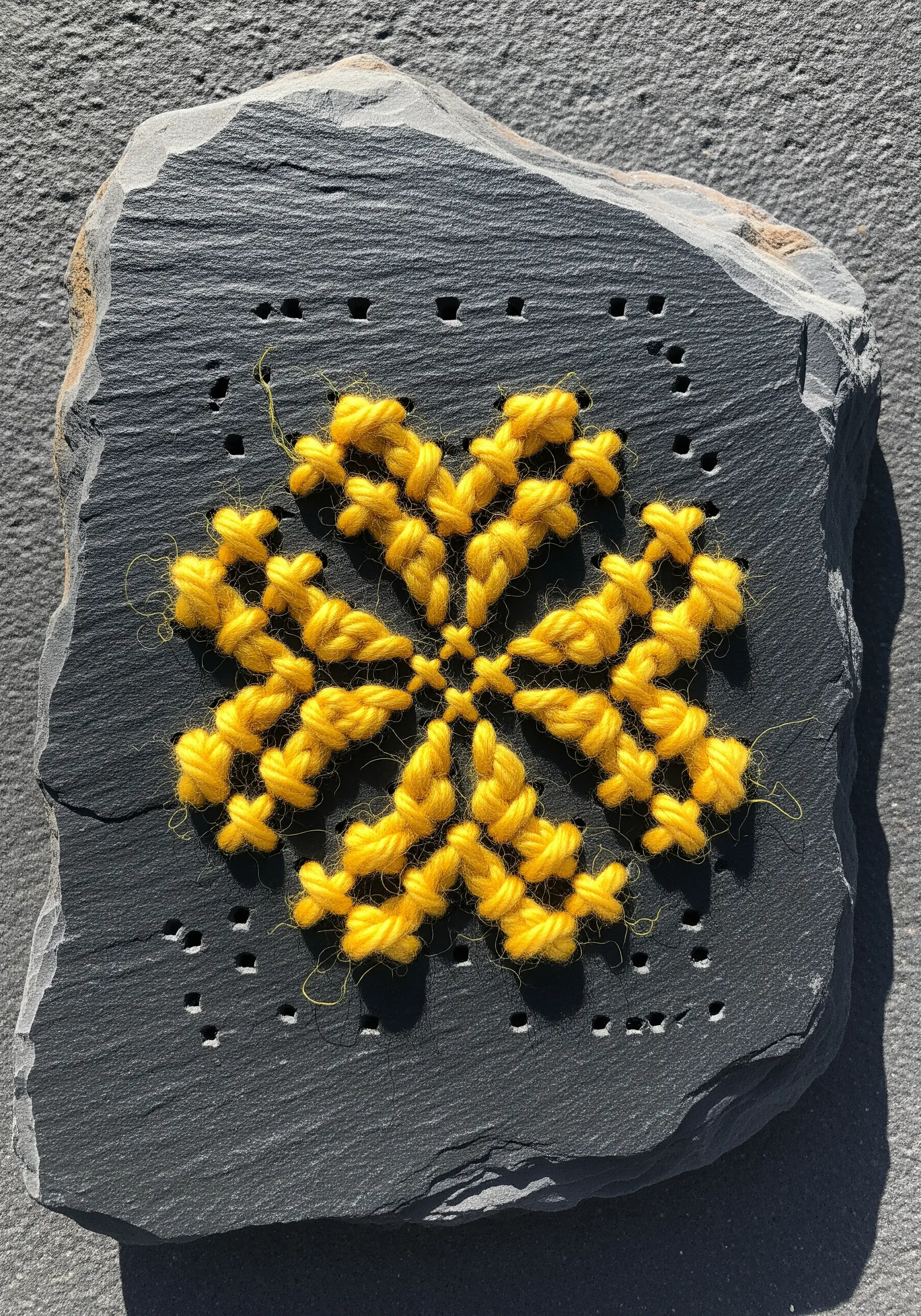 Slate stone with drilled holes and yellow yarn cross-stitch pattern