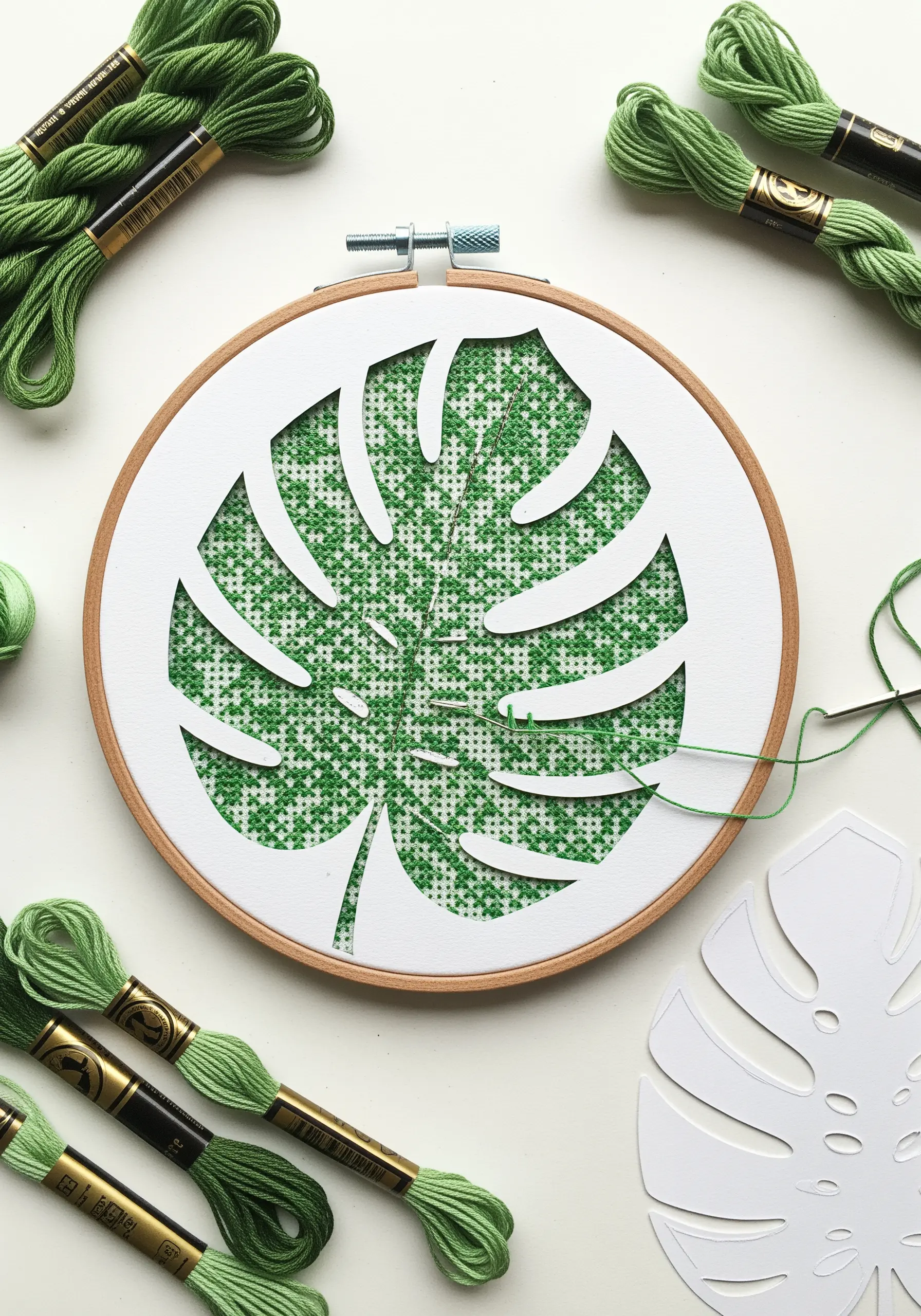 A paper-cut monstera leaf stencil placed over Aida cloth, being filled in with green cross-stitches.