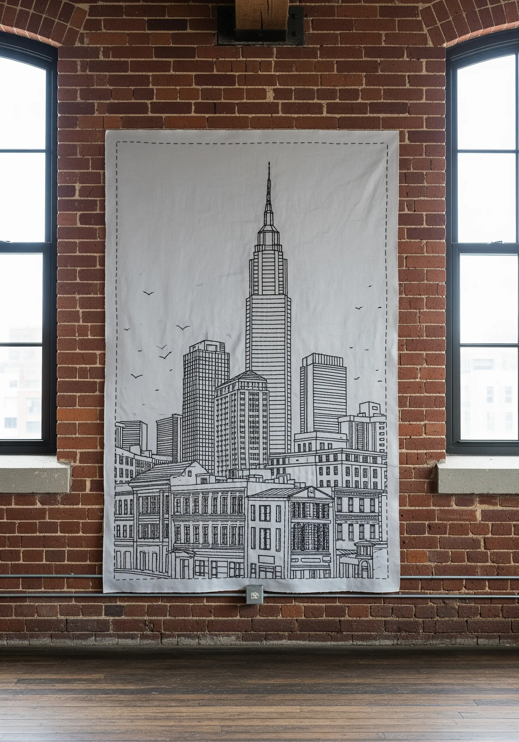 A large, white fabric panel with a black line-art embroidery of a city skyline against a brick wall.