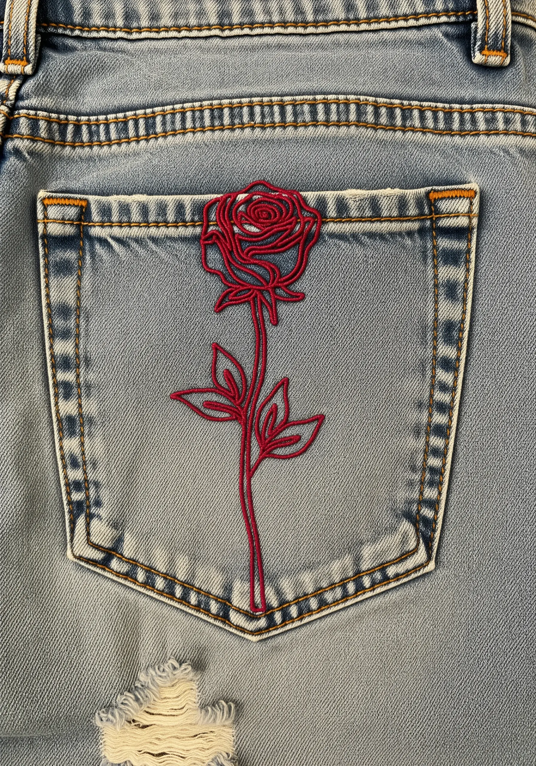 A stylized red rose embroidered on the back pocket of light-wash denim jeans.