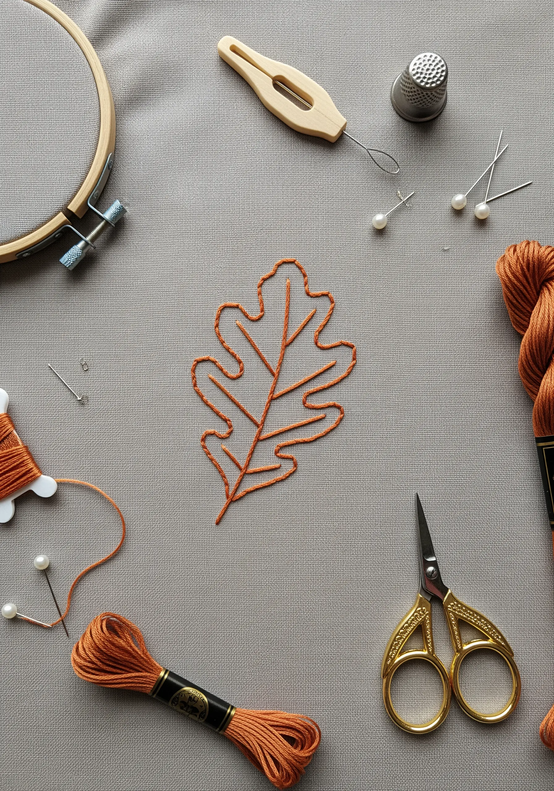 A simple outline of an oak leaf in burnt orange thread on gray fabric.