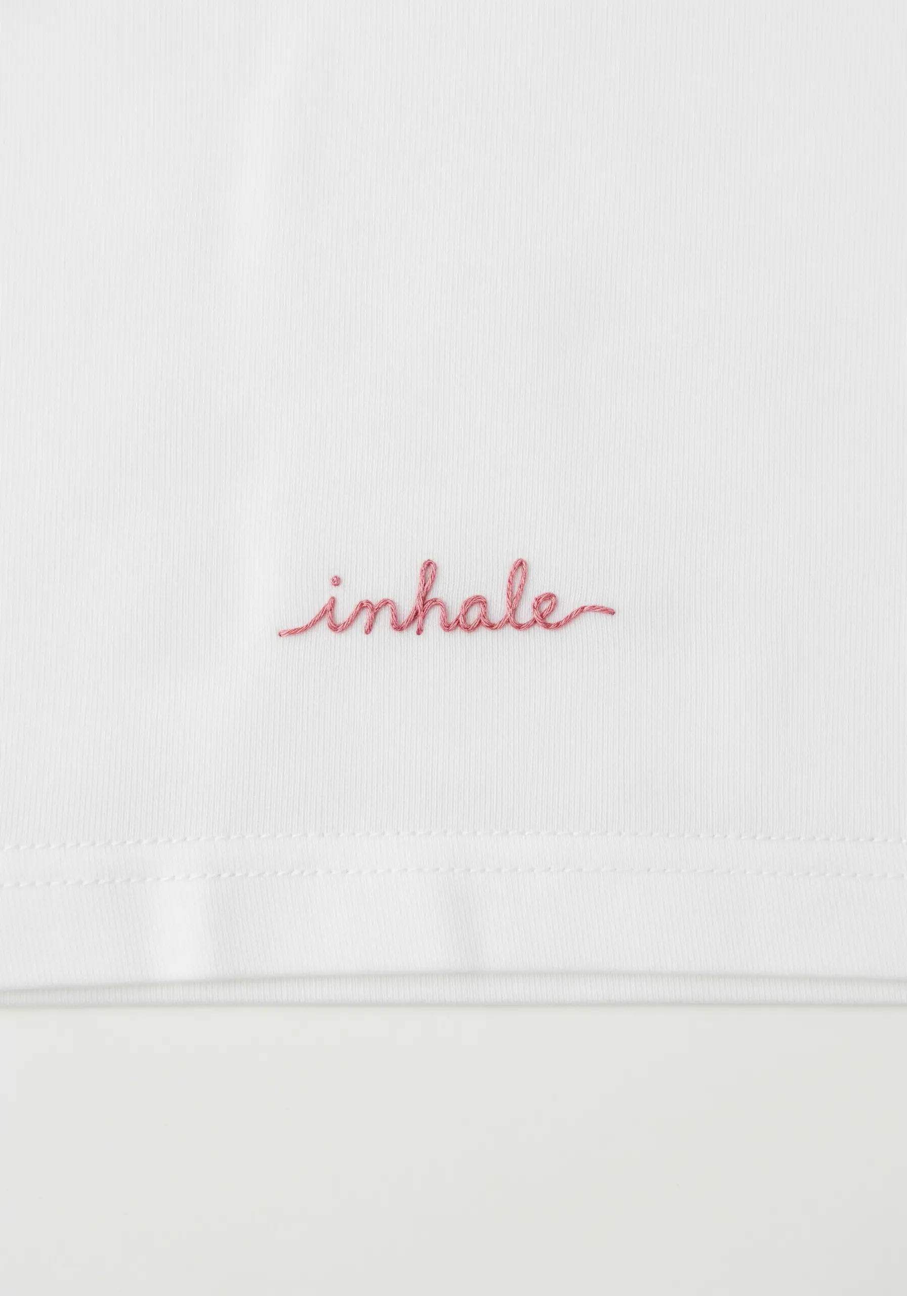 The word 'inhale' embroidered in delicate pink script on a white t-shirt.