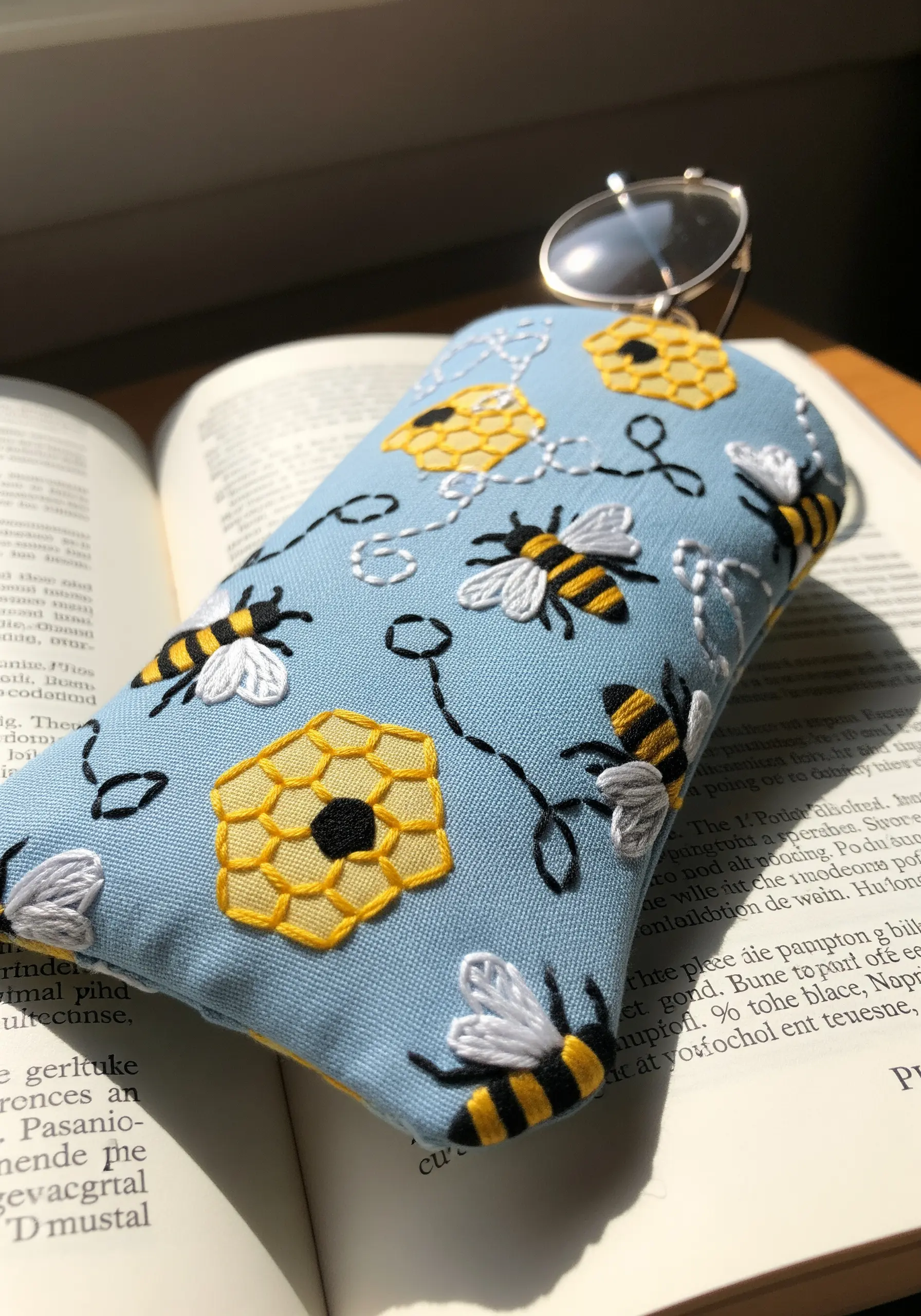 A light blue padded eyeglass case embroidered with a playful pattern of bees and honeycombs.