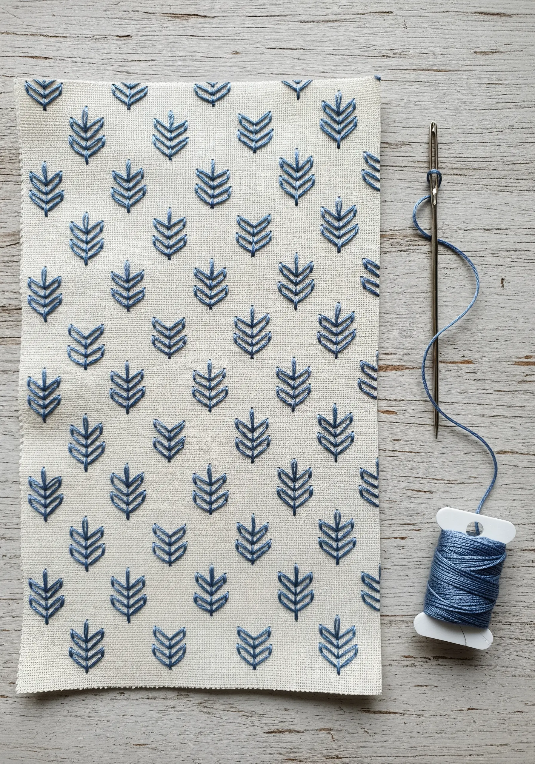 A repeating pattern of small, geometric blue leaf motifs embroidered on off-white fabric.