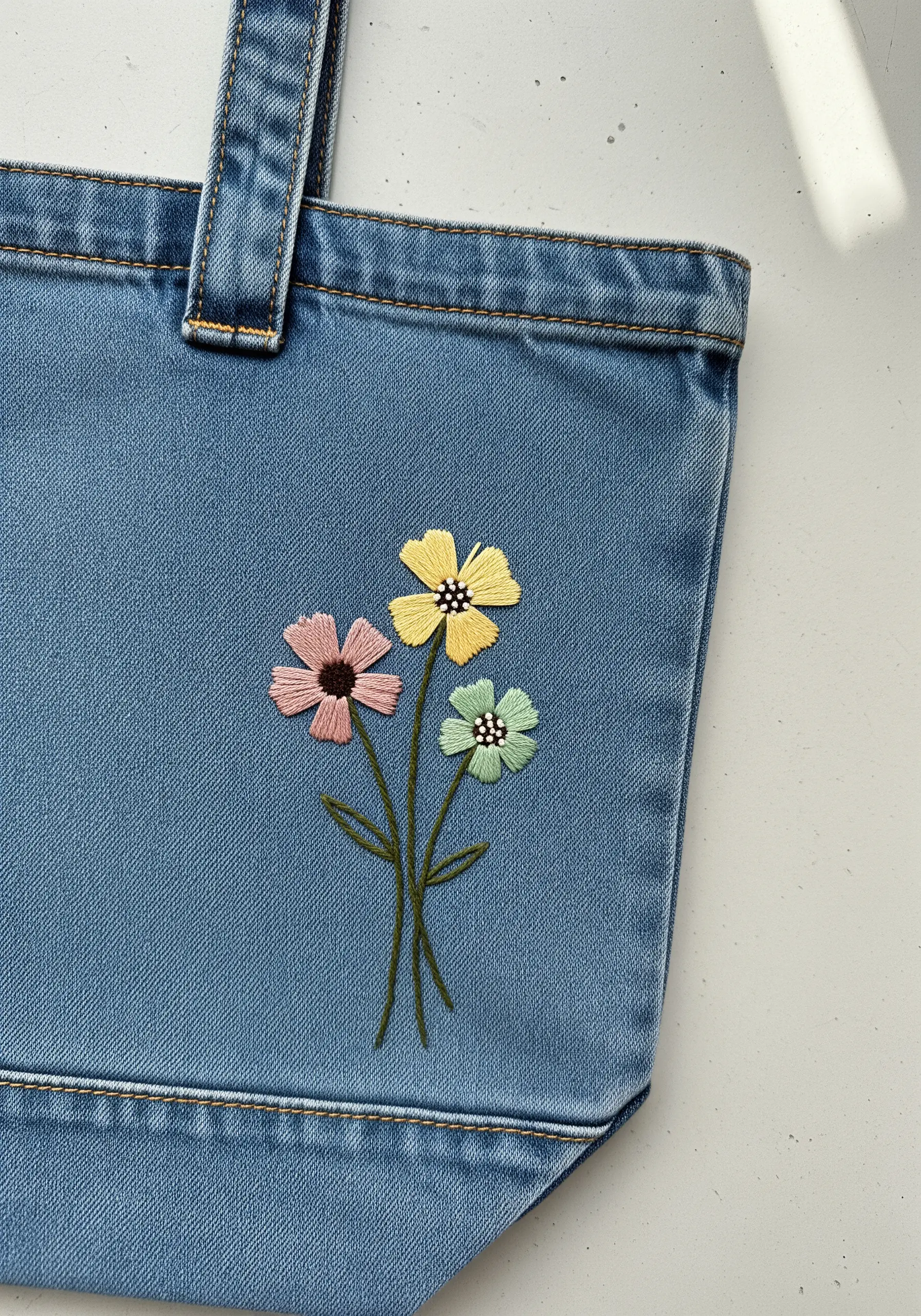 A small bouquet of three pastel-colored flowers embroidered on a denim tote bag.