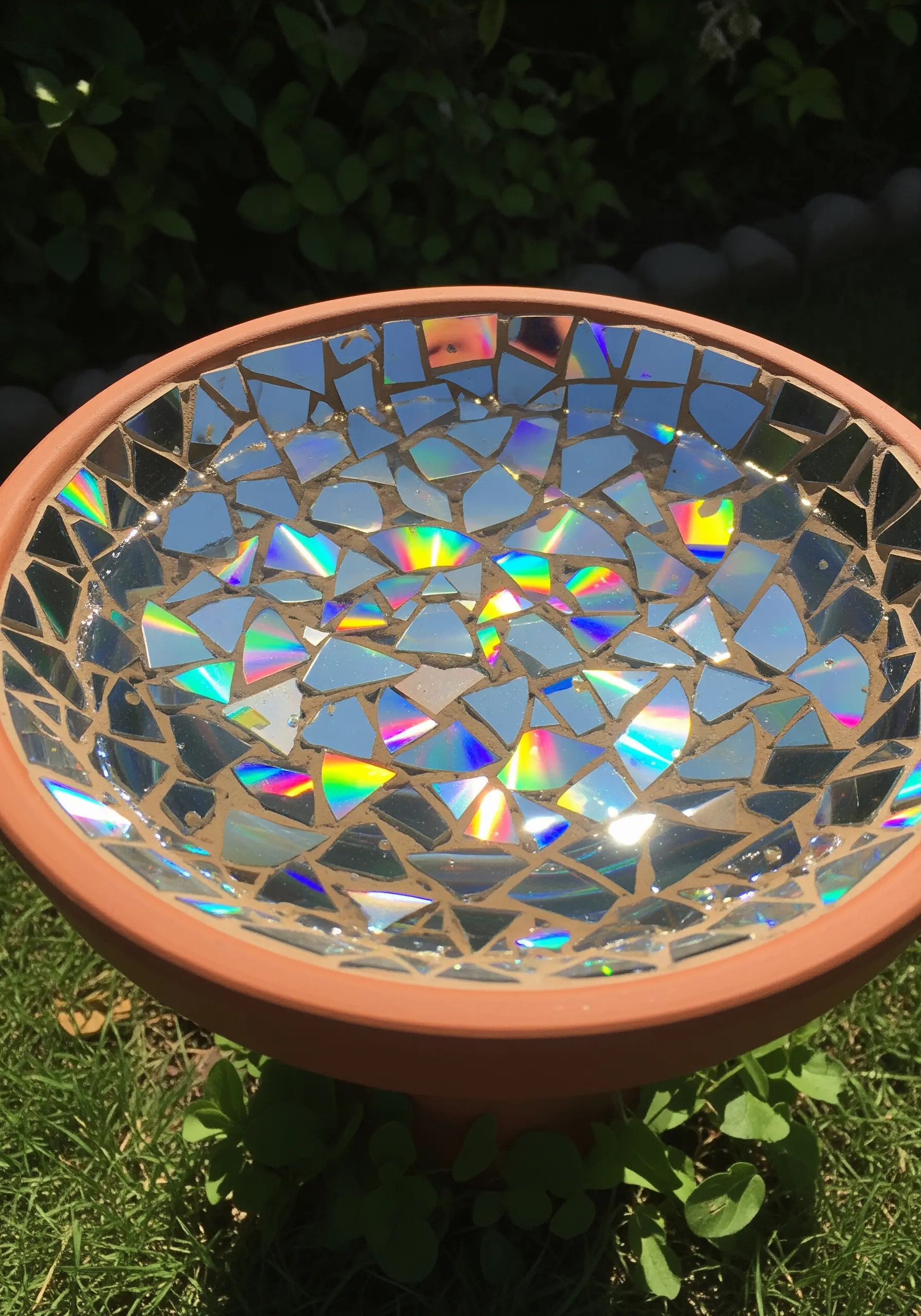 A terracotta bird bath with its basin covered in a mosaic of iridescent, shattered CD pieces.