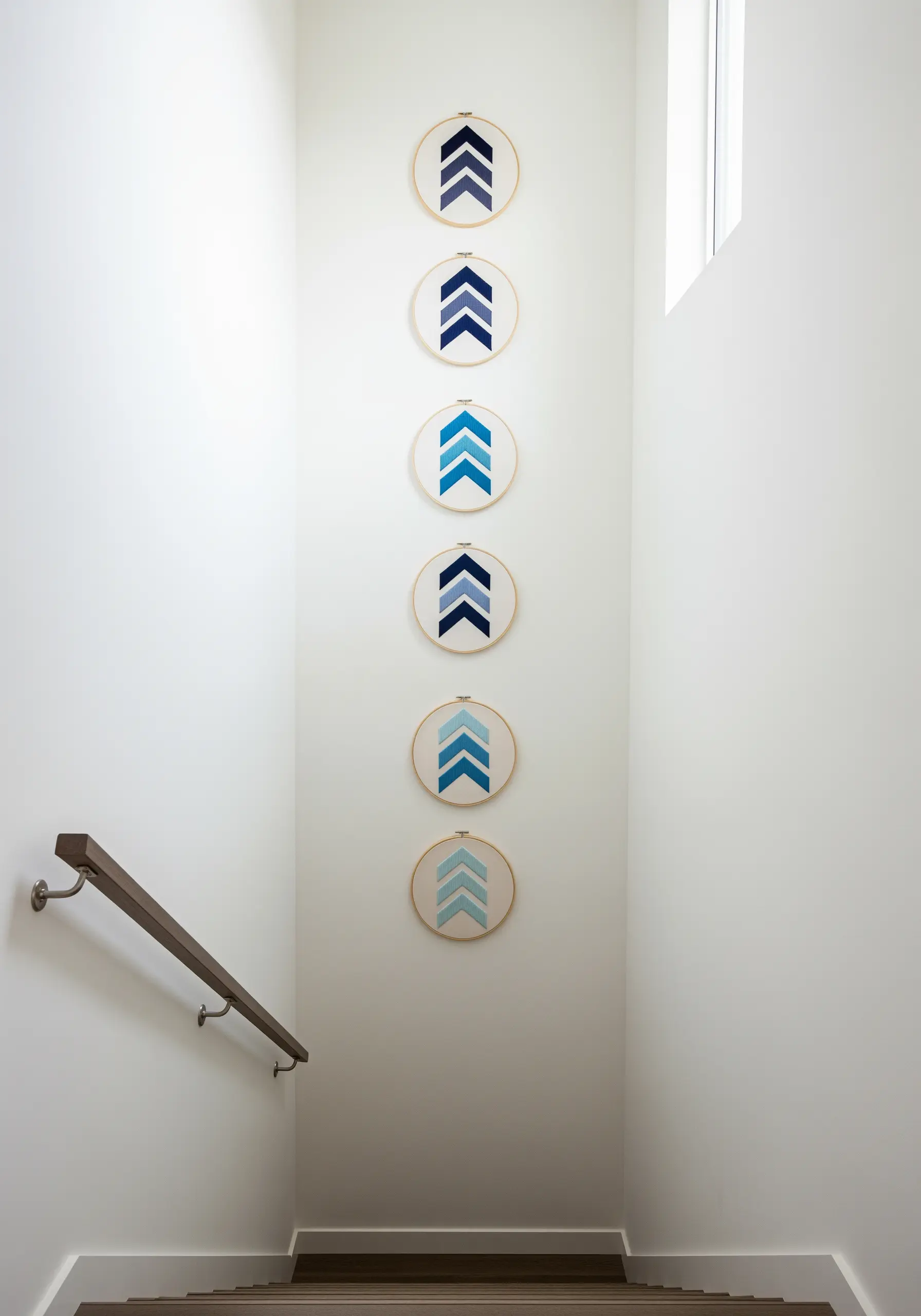 Six embroidery hoops with blue gradient chevron patterns hung vertically on a tall staircase wall.