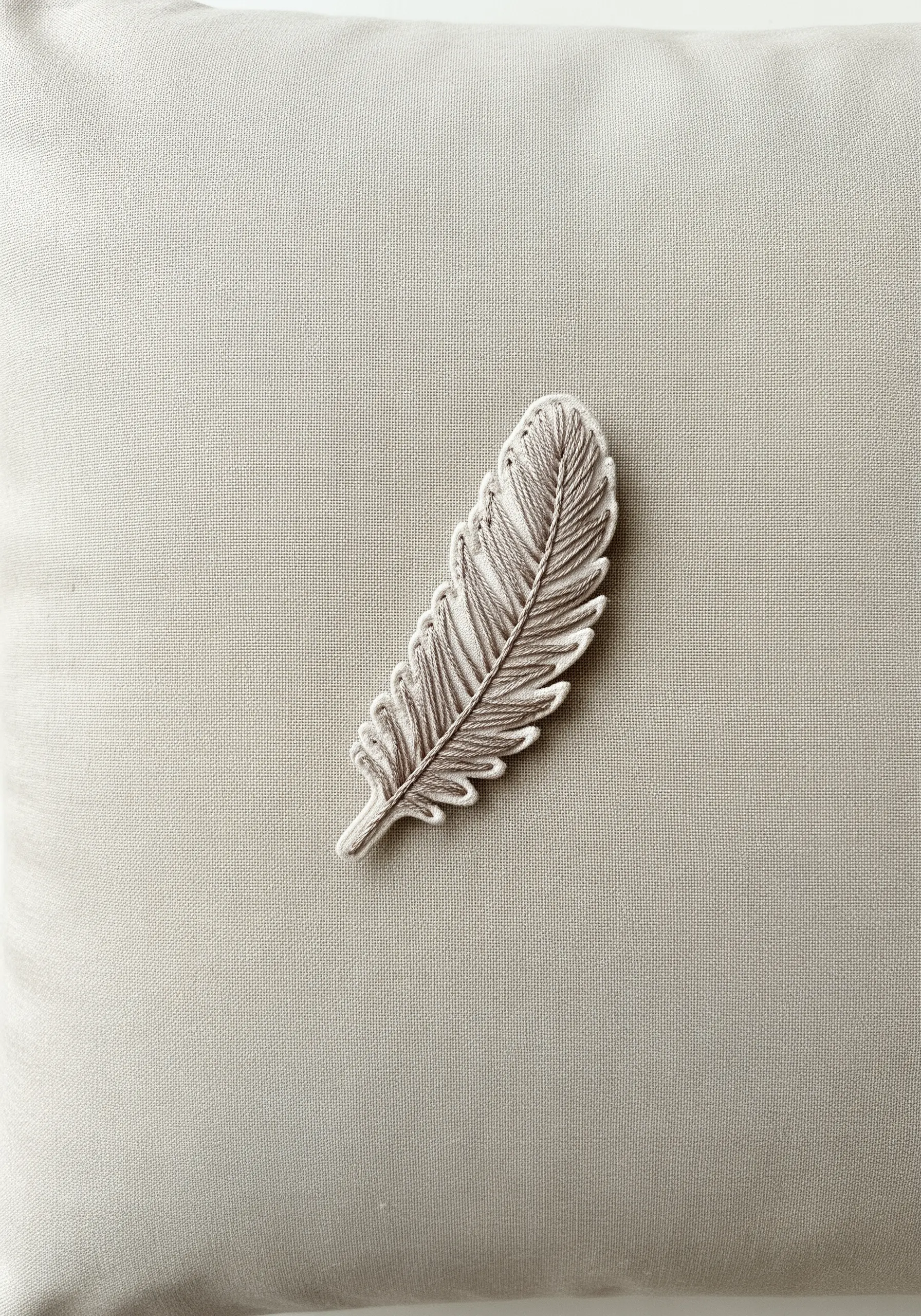 A single, detailed feather embroidered in shades of gray and white on a light beige pillowcase.