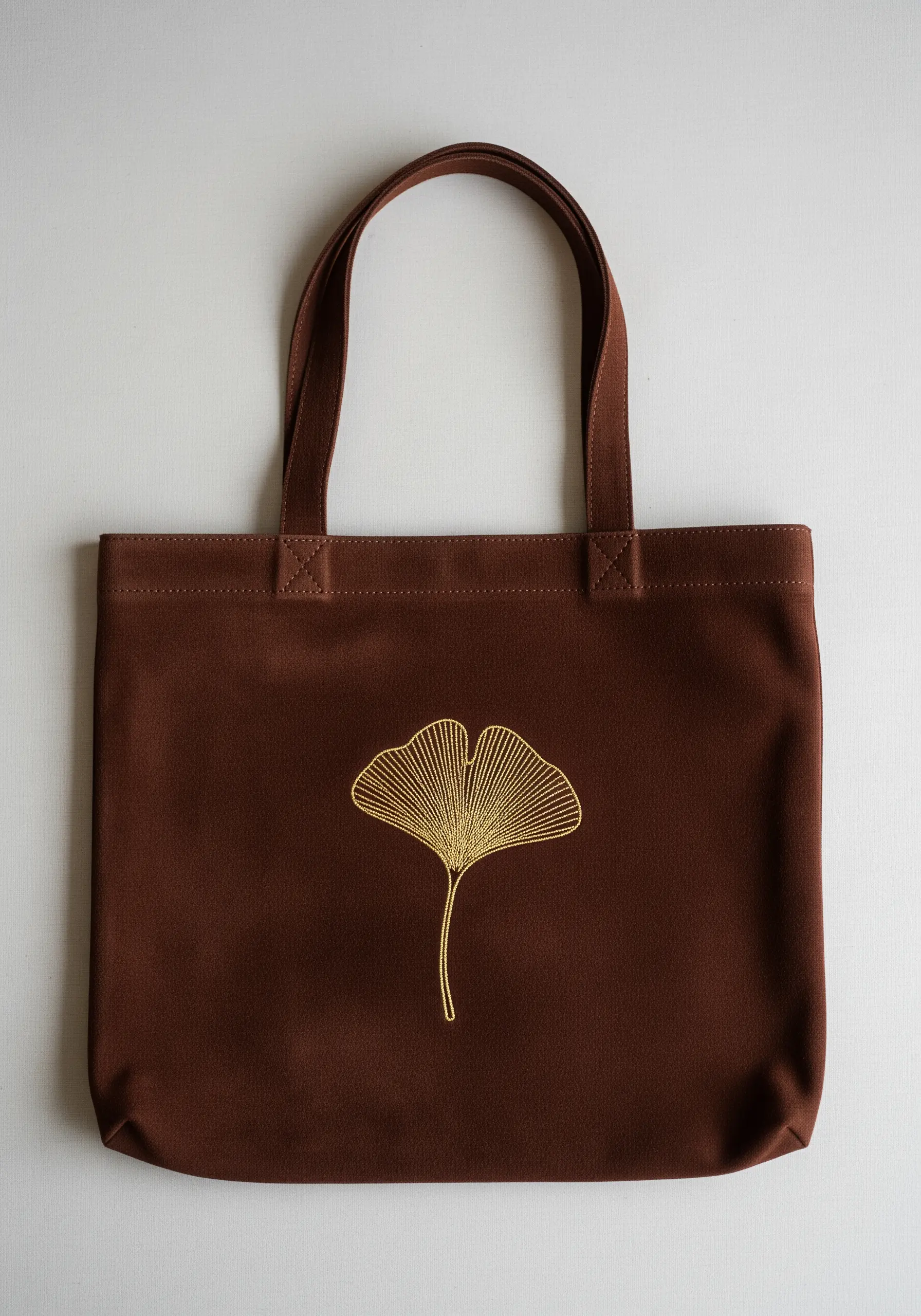 A single ginkgo leaf embroidered in gold thread on a dark brown canvas tote bag.