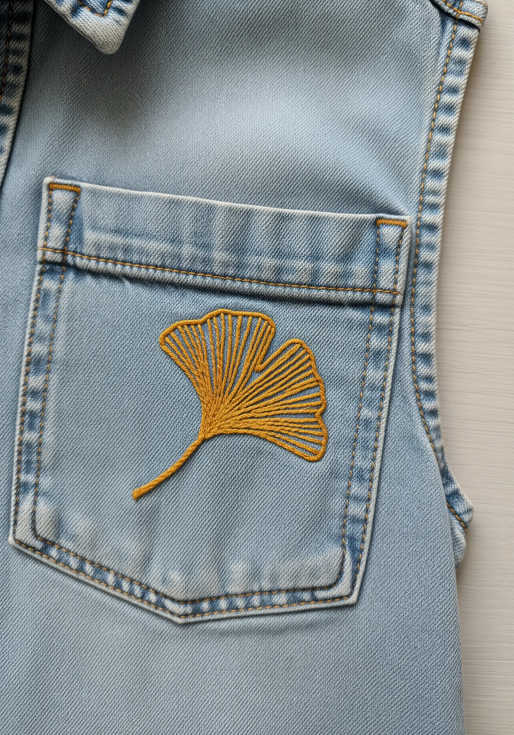A yellow satin-stitched ginkgo leaf embroidered on the pocket of a denim vest.