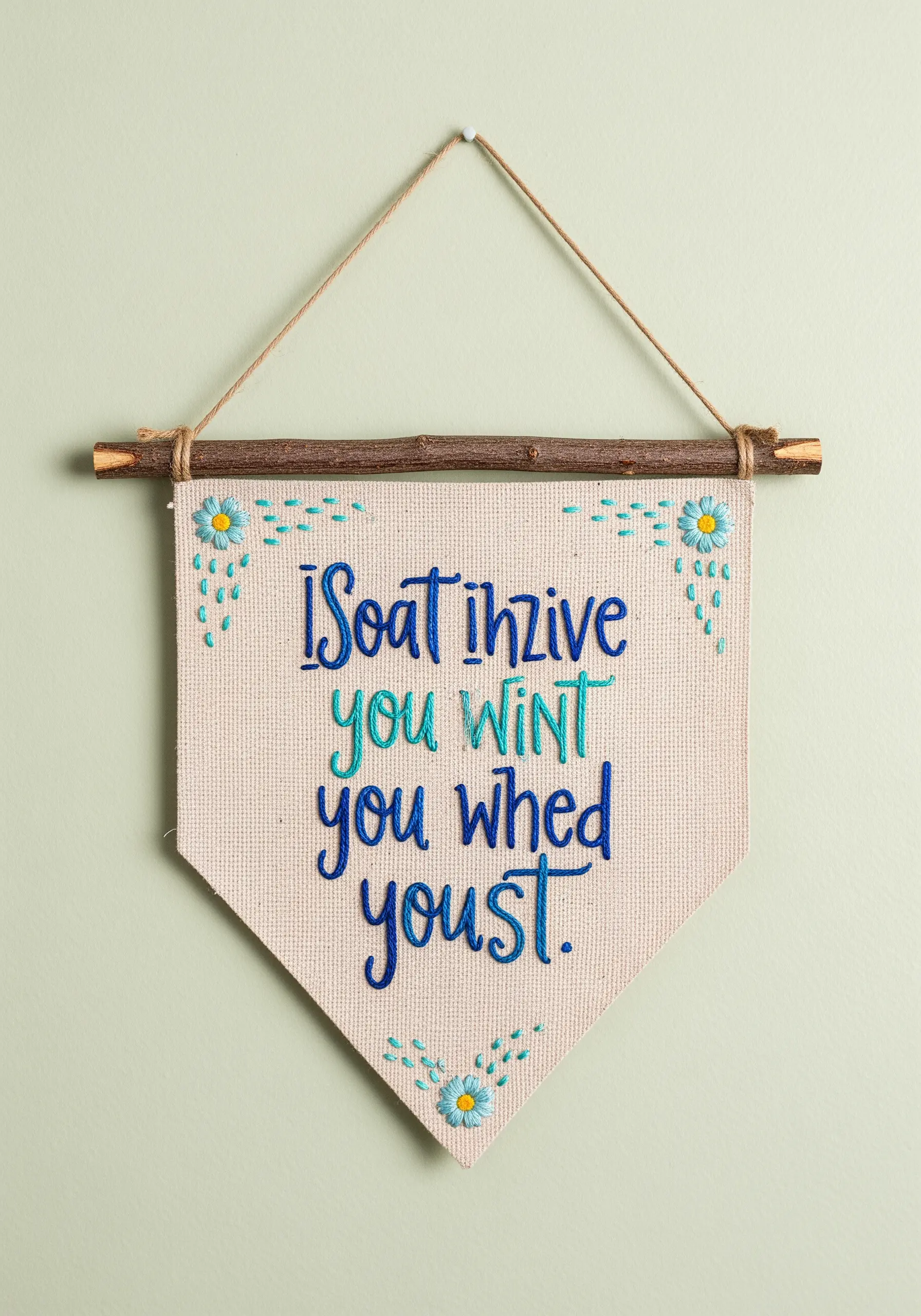 Embroidered quote on a canvas pennant hanging from a wooden stick on a light green wall.
