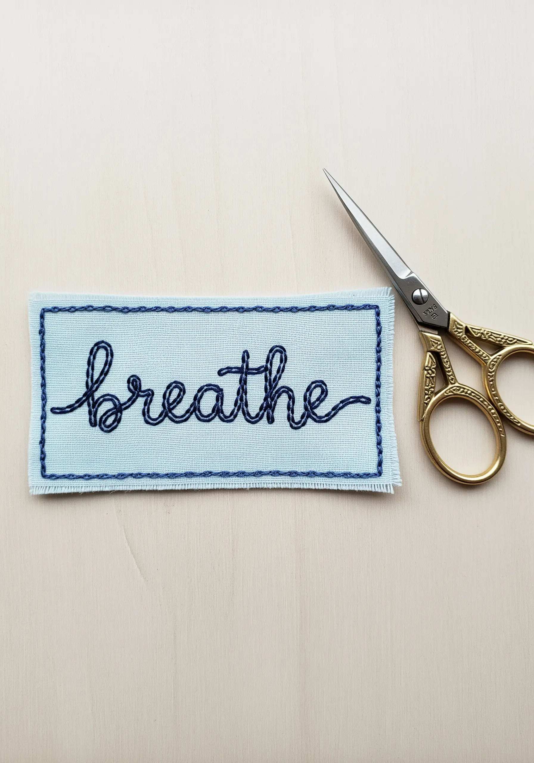 The word 'breathe' embroidered in cursive on a light blue fabric patch next to scissors.