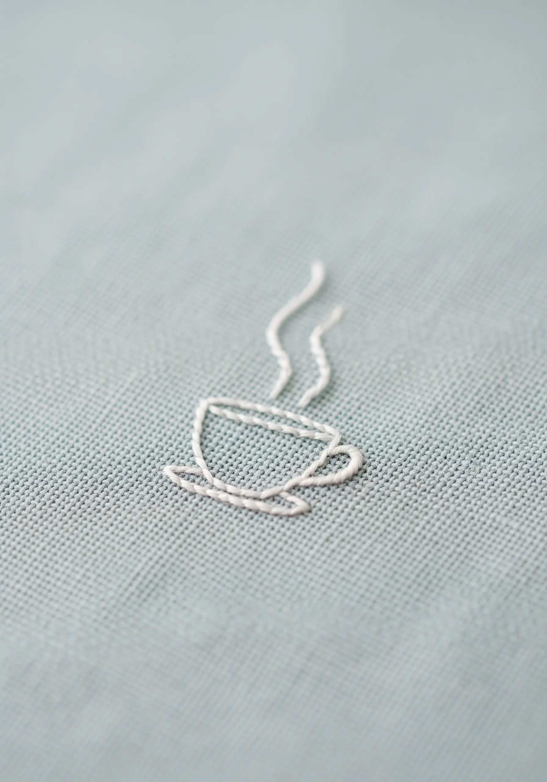 A minimalist line art coffee cup stitched in white thread on a muted blue fabric.