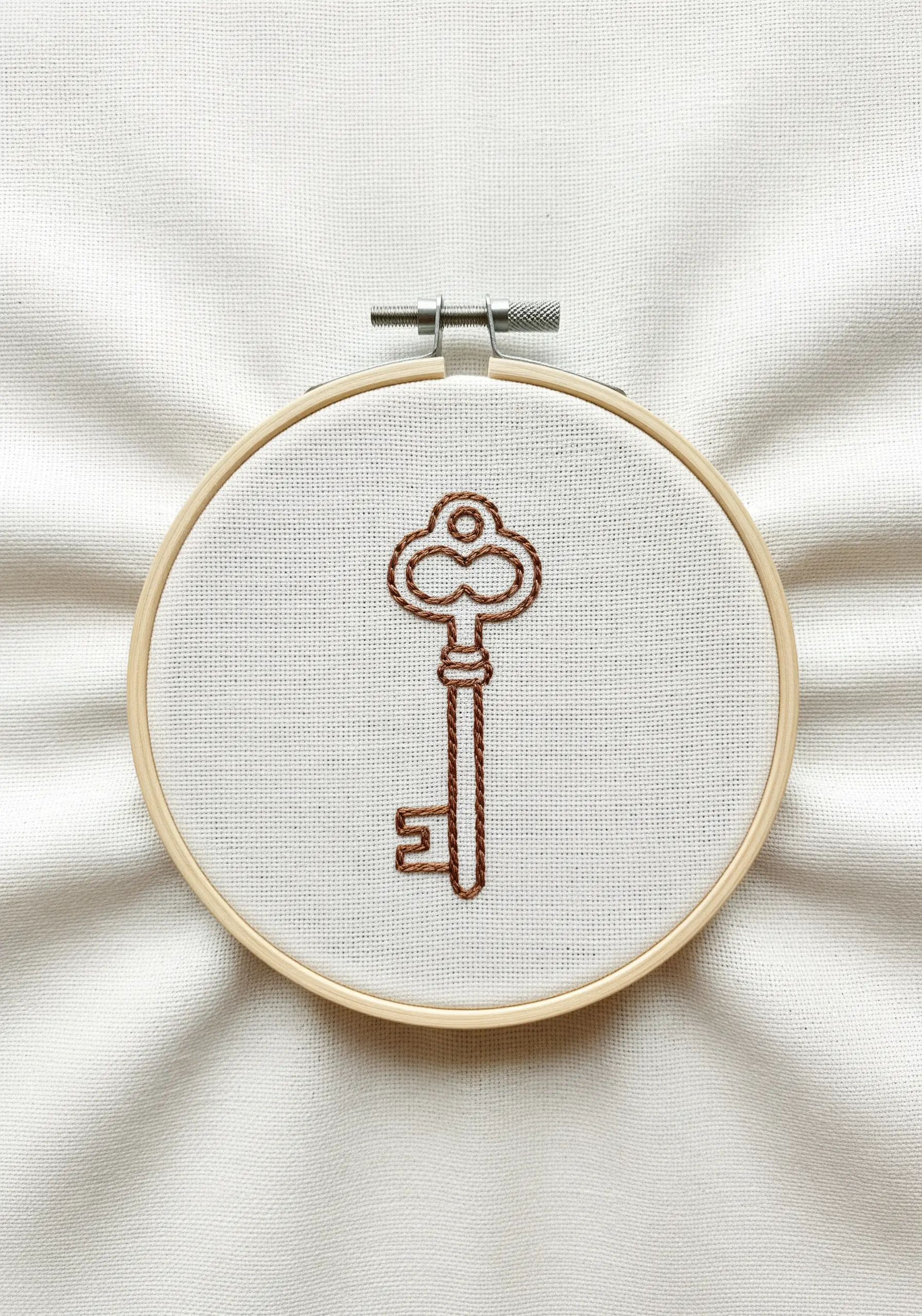 An outline of an antique key stitched in brown thread on white fabric.