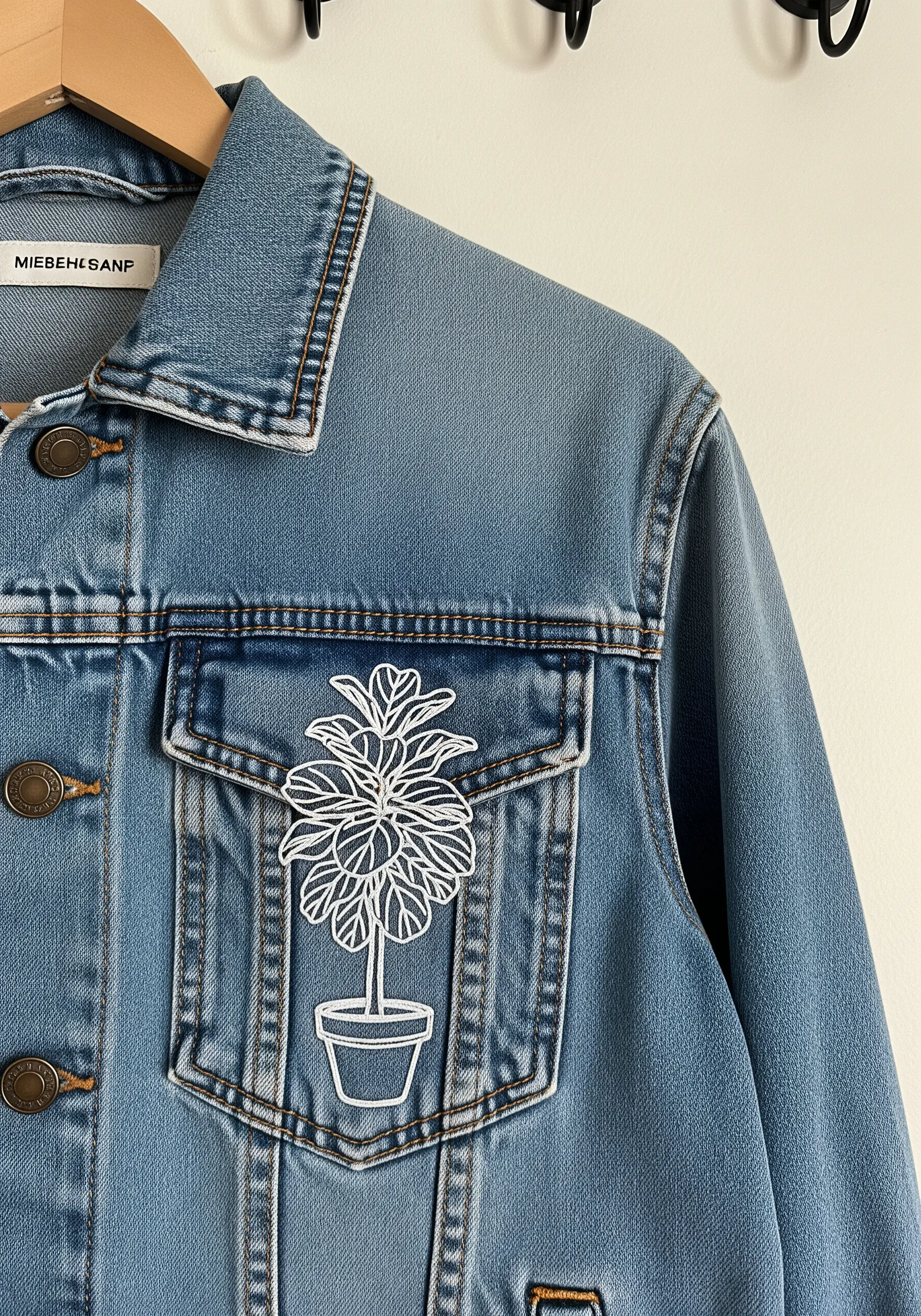 A white line-art embroidery of a potted fiddle leaf fig plant on a denim jacket pocket.