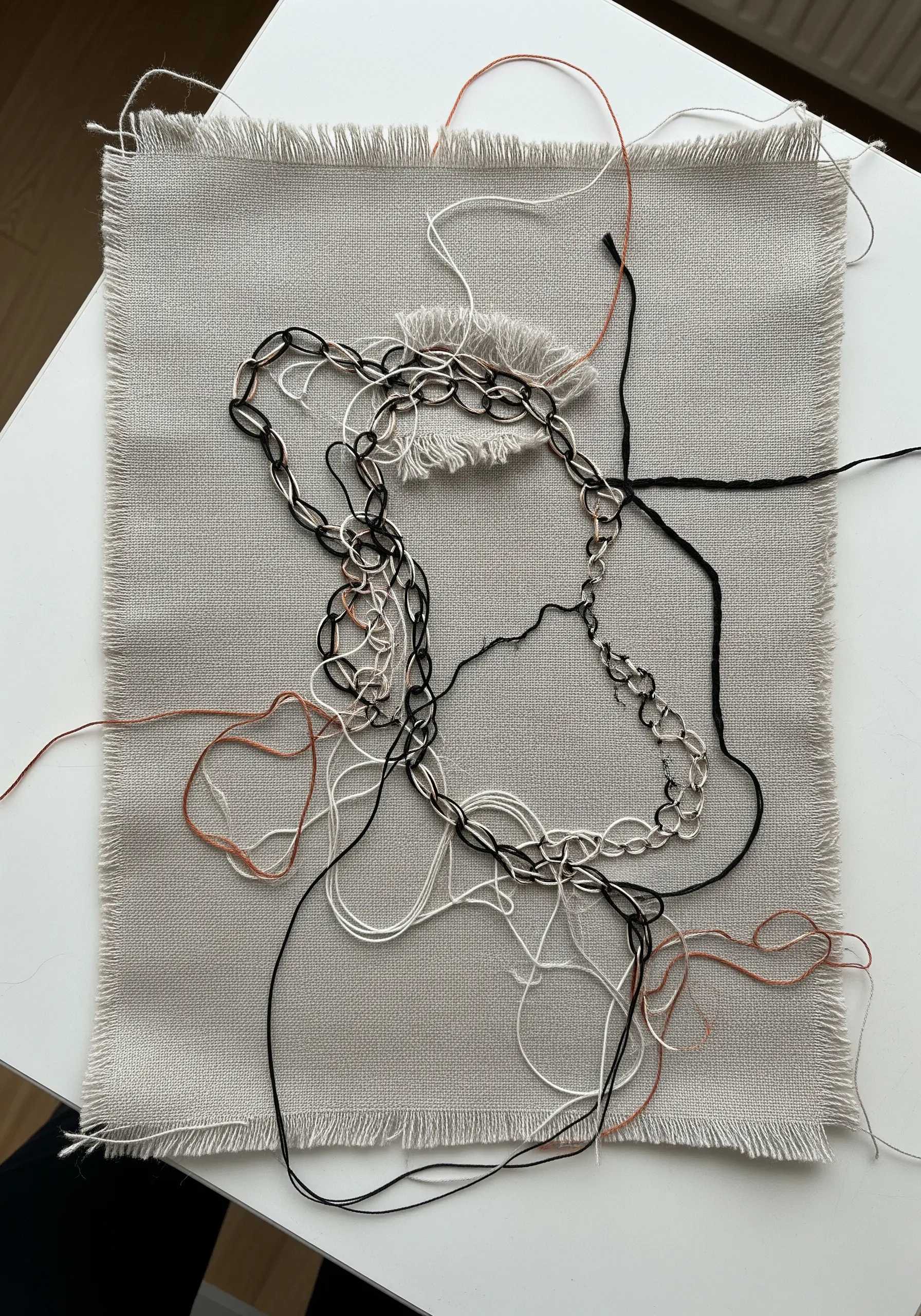 A deconstructed embroidery piece on raw-edge linen with loose threads and metal chains.
