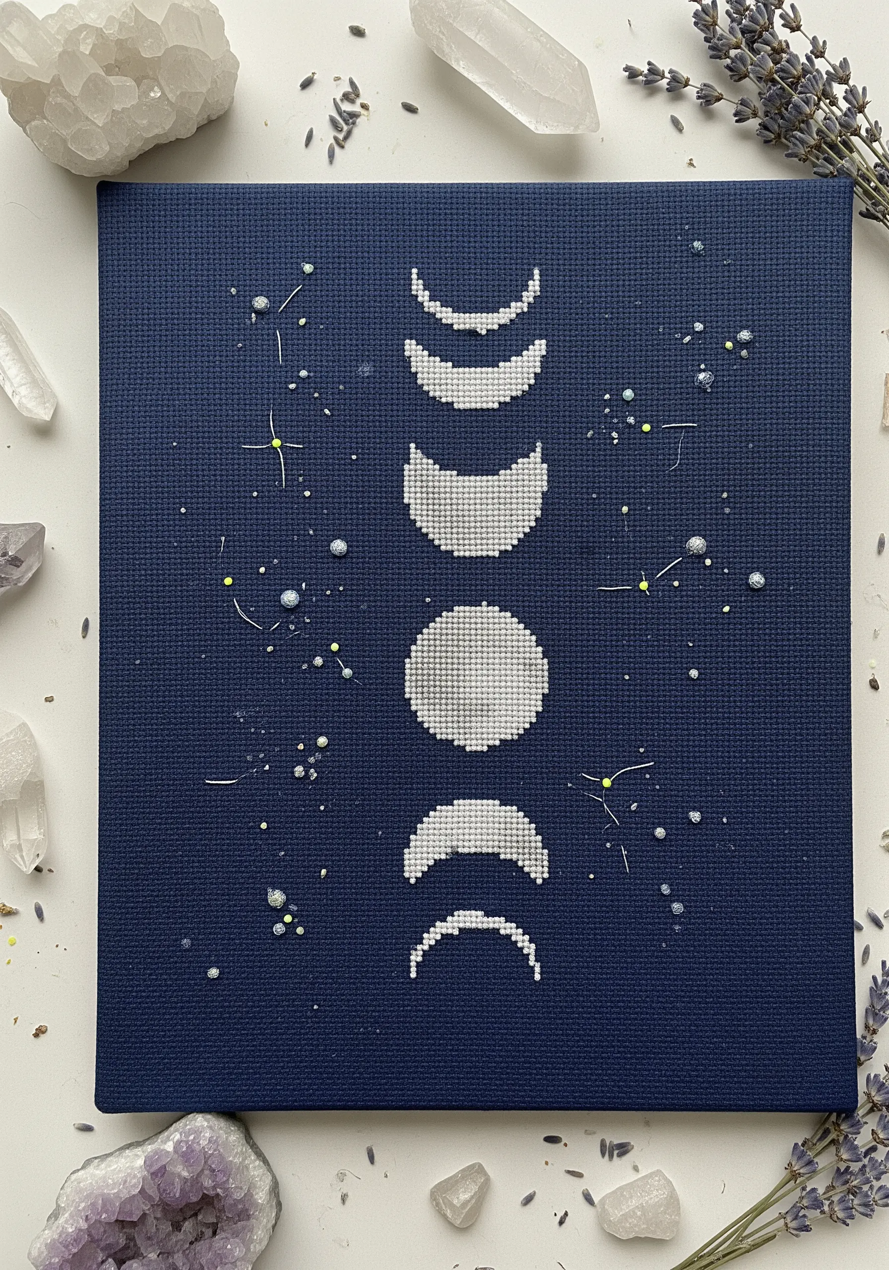 Minimalist cross-stitch of moon phases in white thread on navy blue Aida fabric.