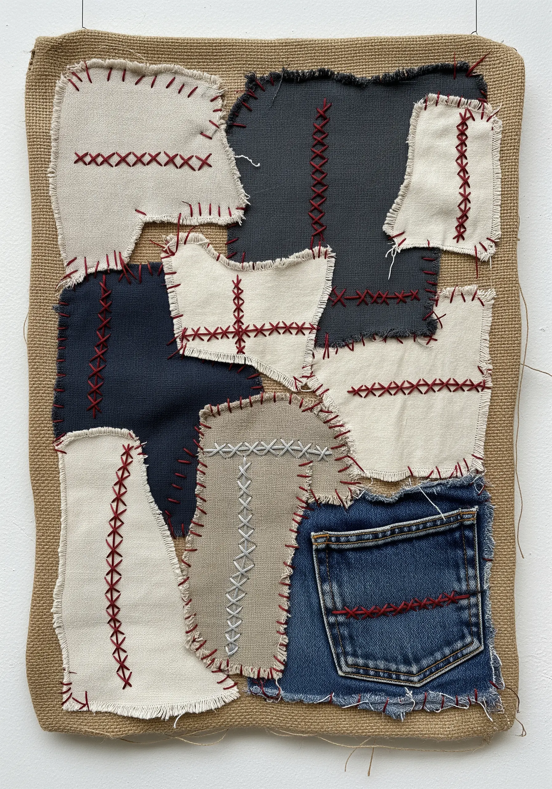 A patchwork collage of neutral and denim fabric scraps stitched together with red thread on burlap.