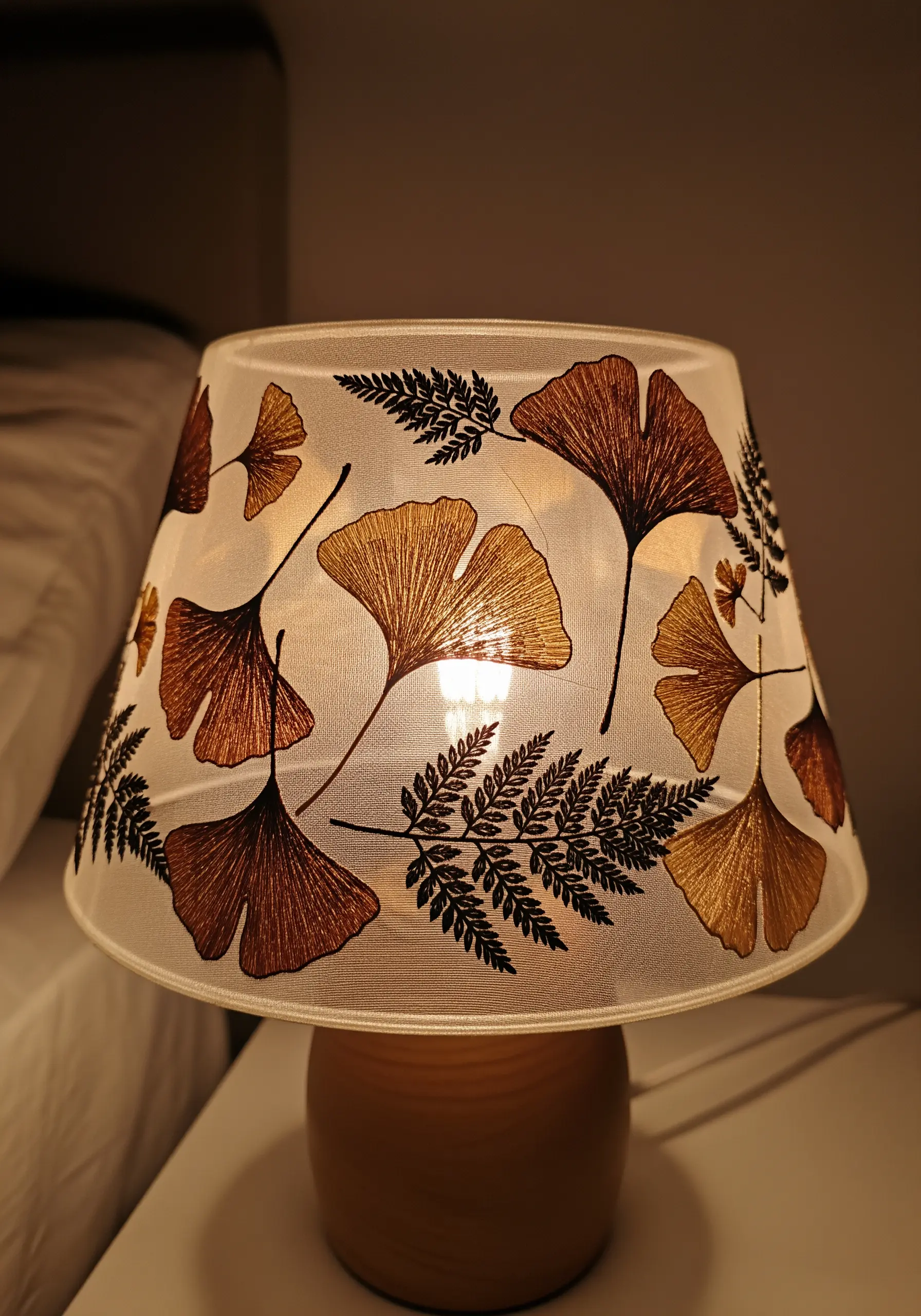 A lit lampshade decorated with a pattern of pressed ginkgo leaves and ferns.