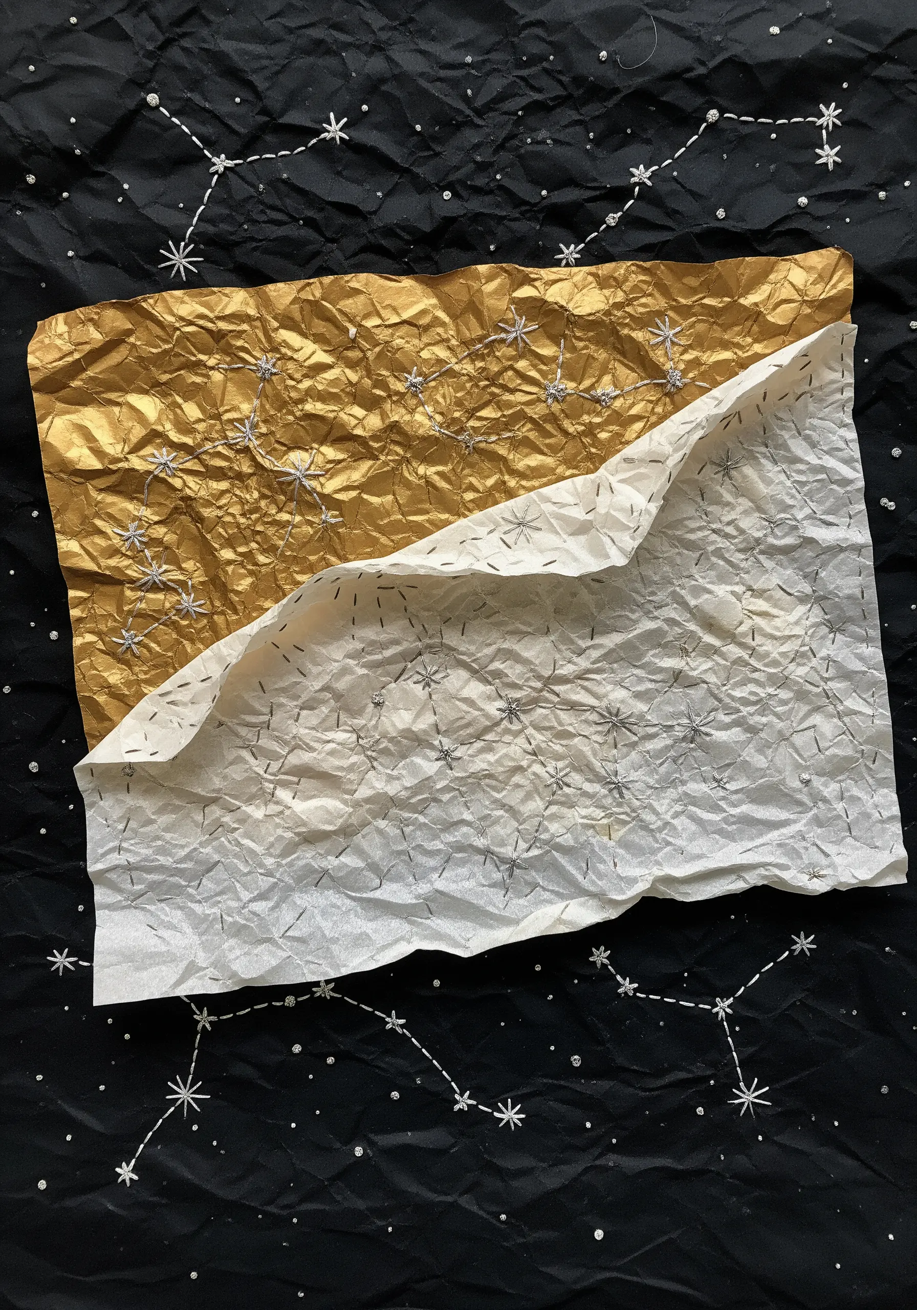 Silver thread constellations stitched onto a crumpled sheet of gold and white paper.