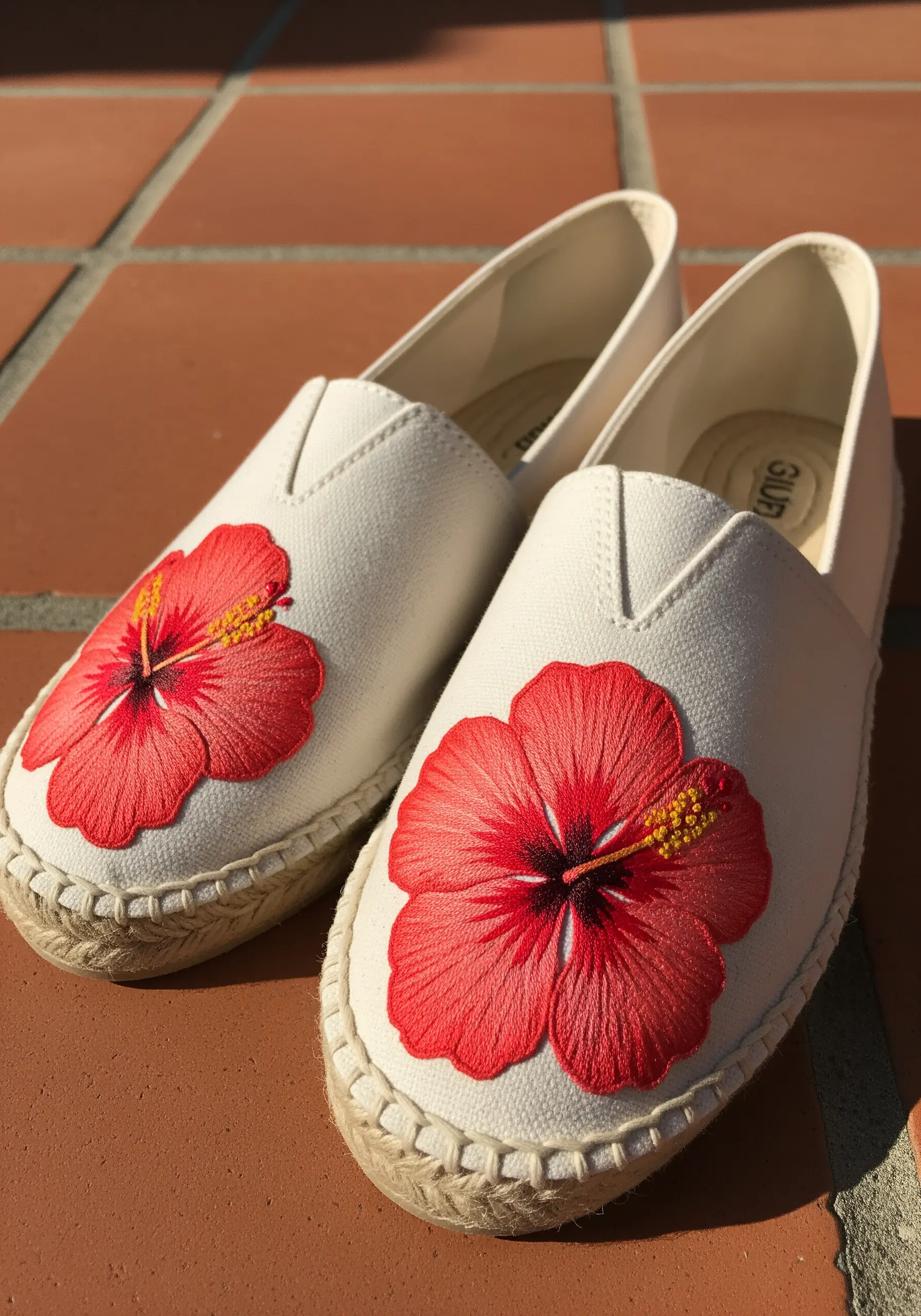 A pair of white canvas espadrilles, each featuring a large, brightly embroidered red hibiscus flower.