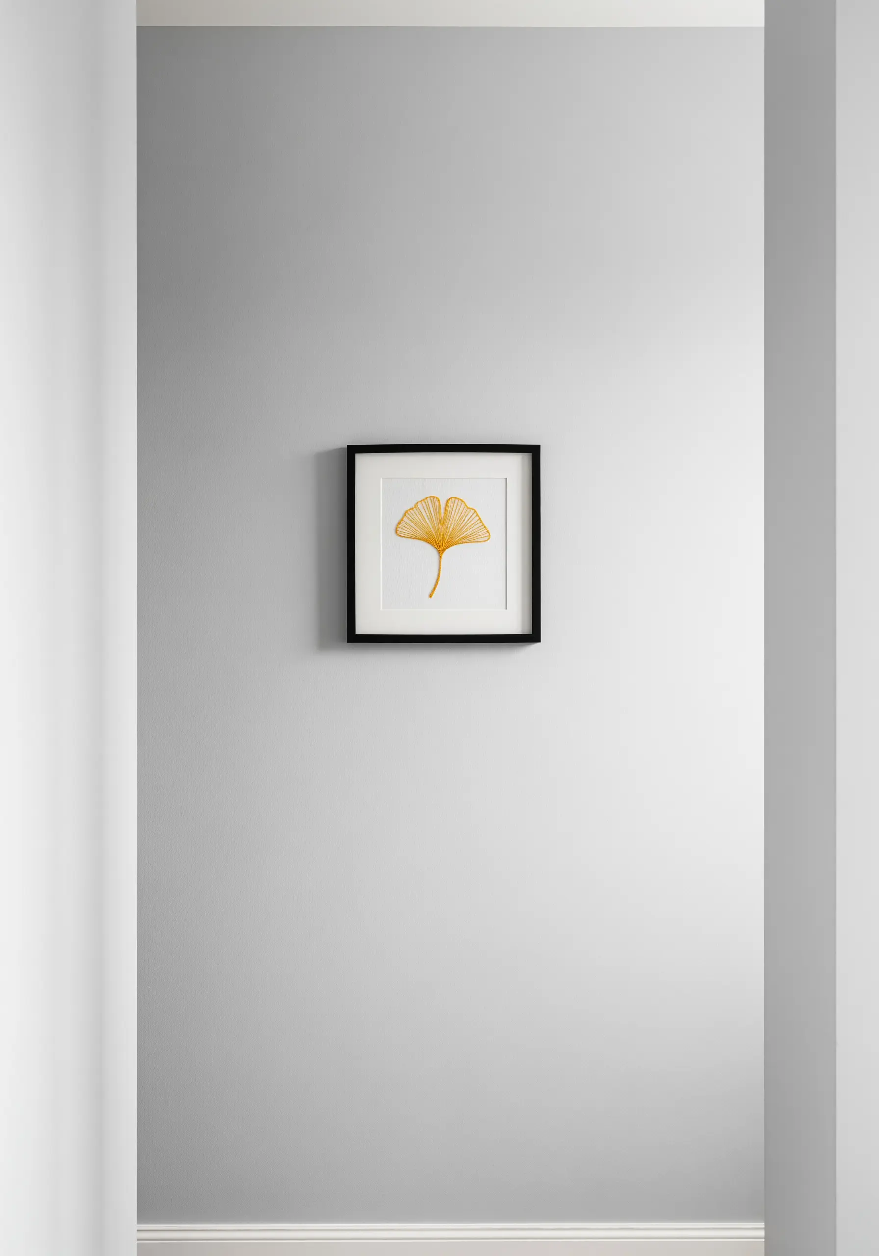 A small, black-framed embroidery of a single yellow ginkgo leaf on a white wall.
