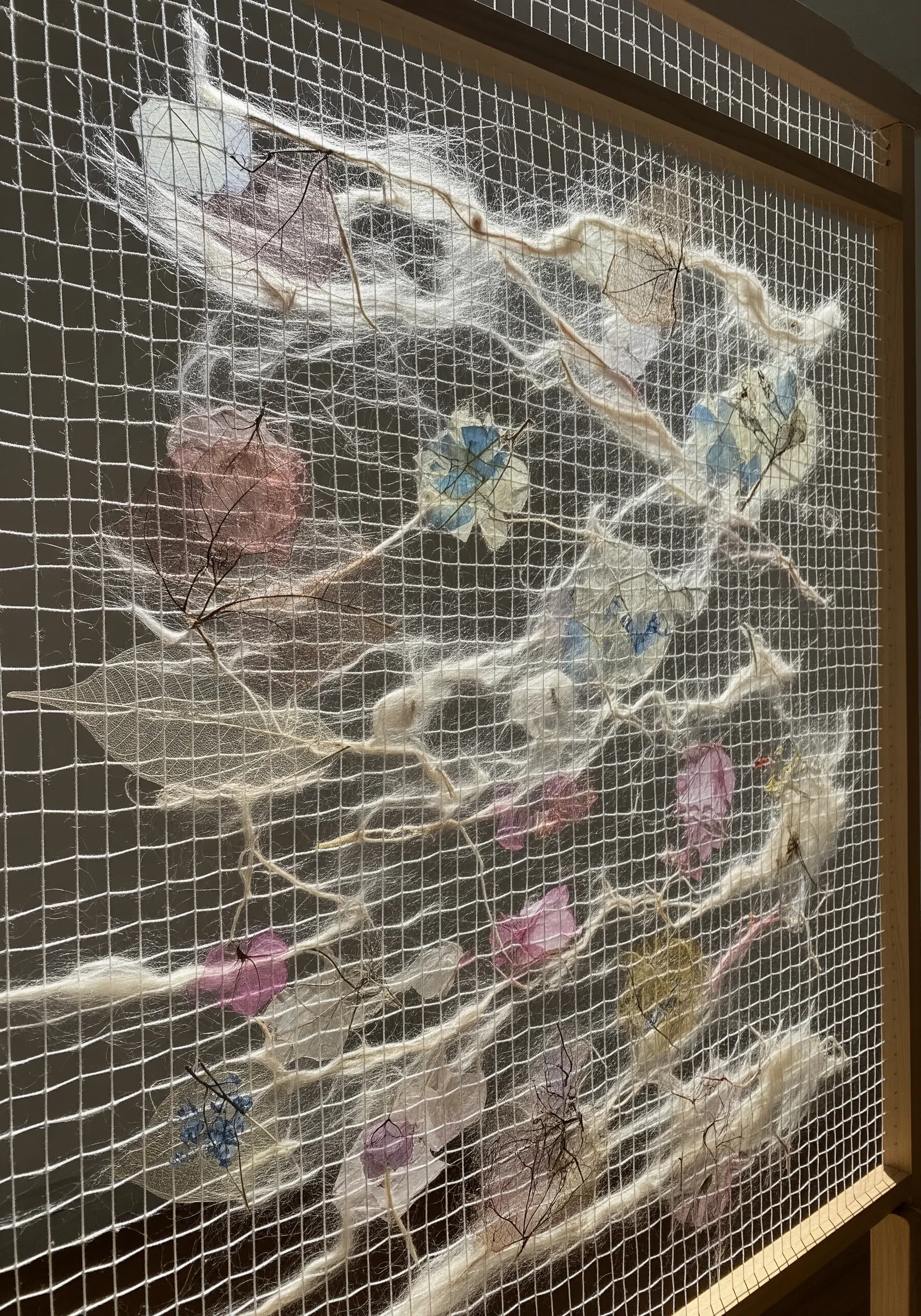 Delicate fiber art on a wire mesh grid, featuring skeleton leaves and wispy white fibers.