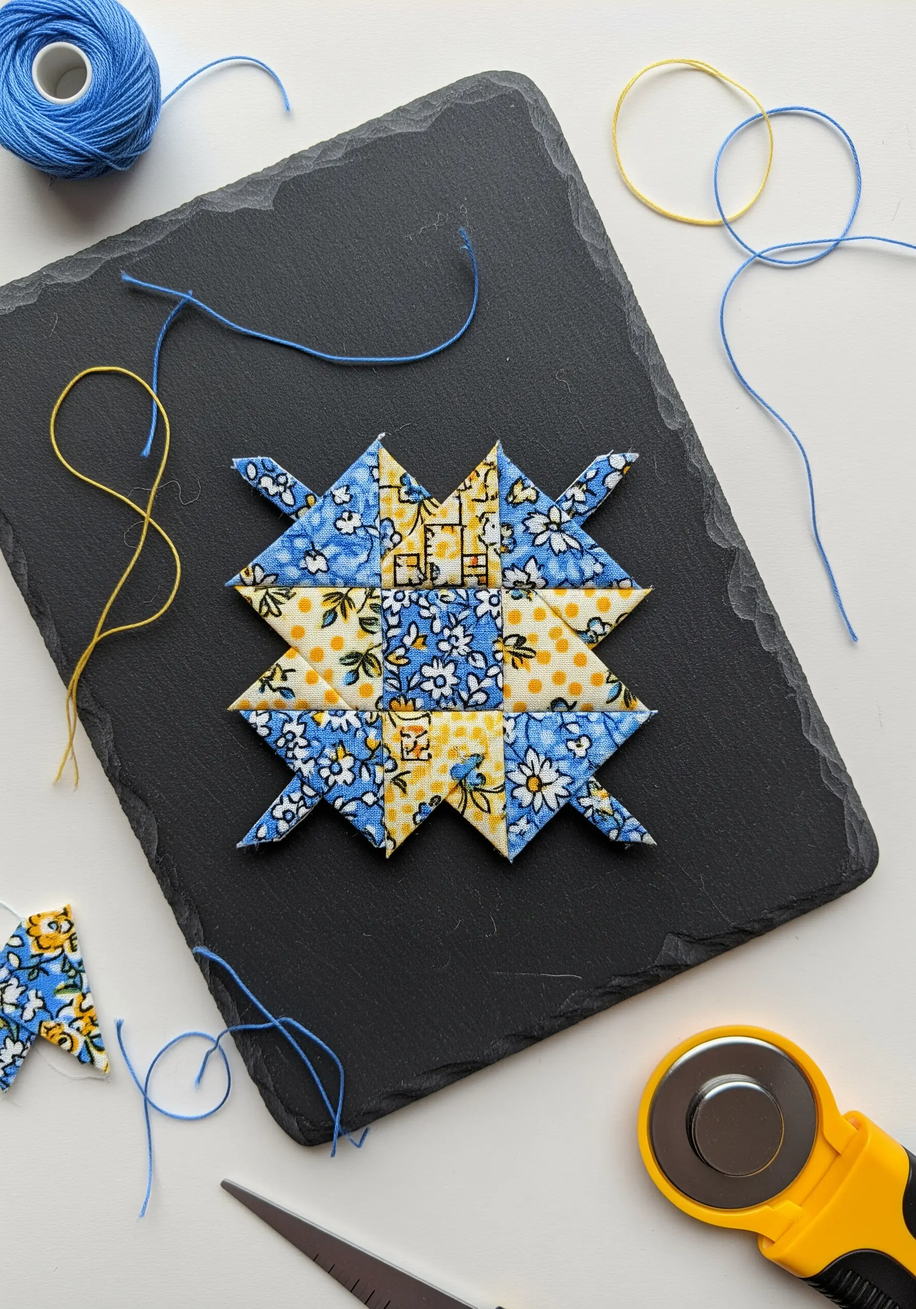 A small, geometric quilted star block made with blue and yellow floral fabrics on a black slate.