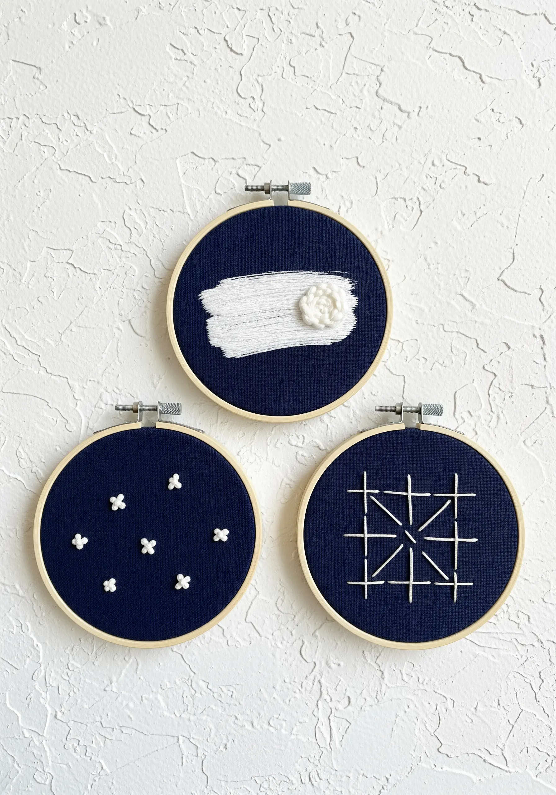 A triptych of minimalist embroidery hoops with white stitches on a dark navy background.