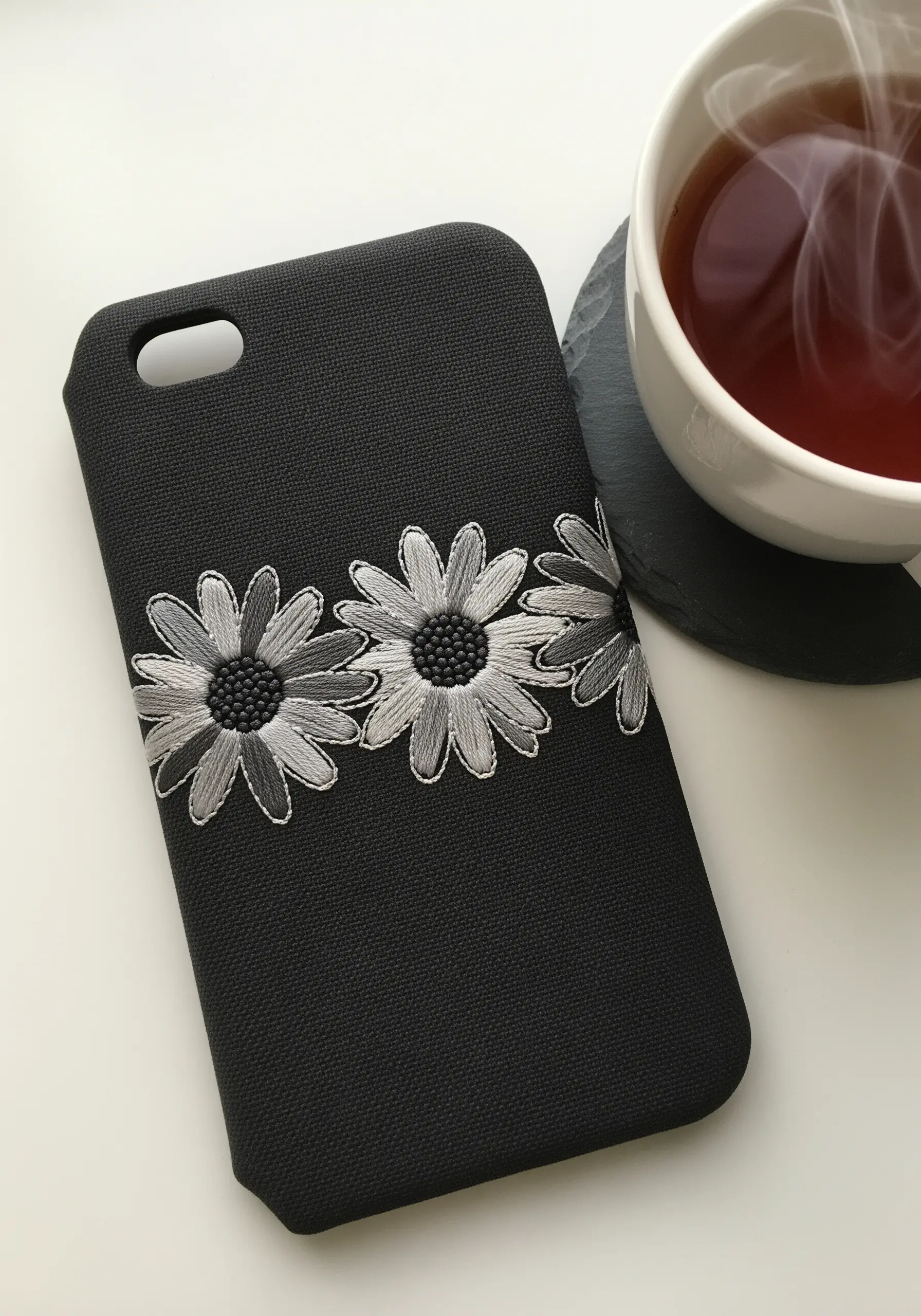 Three daisies embroidered in shades of gray on a black phone case.