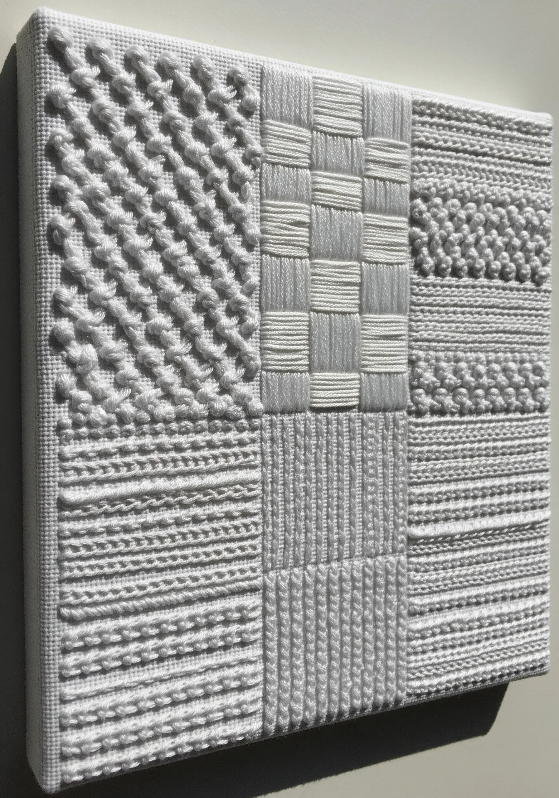 A monochrome white-on-white embroidery sampler with different textural stitch patterns in a grid.