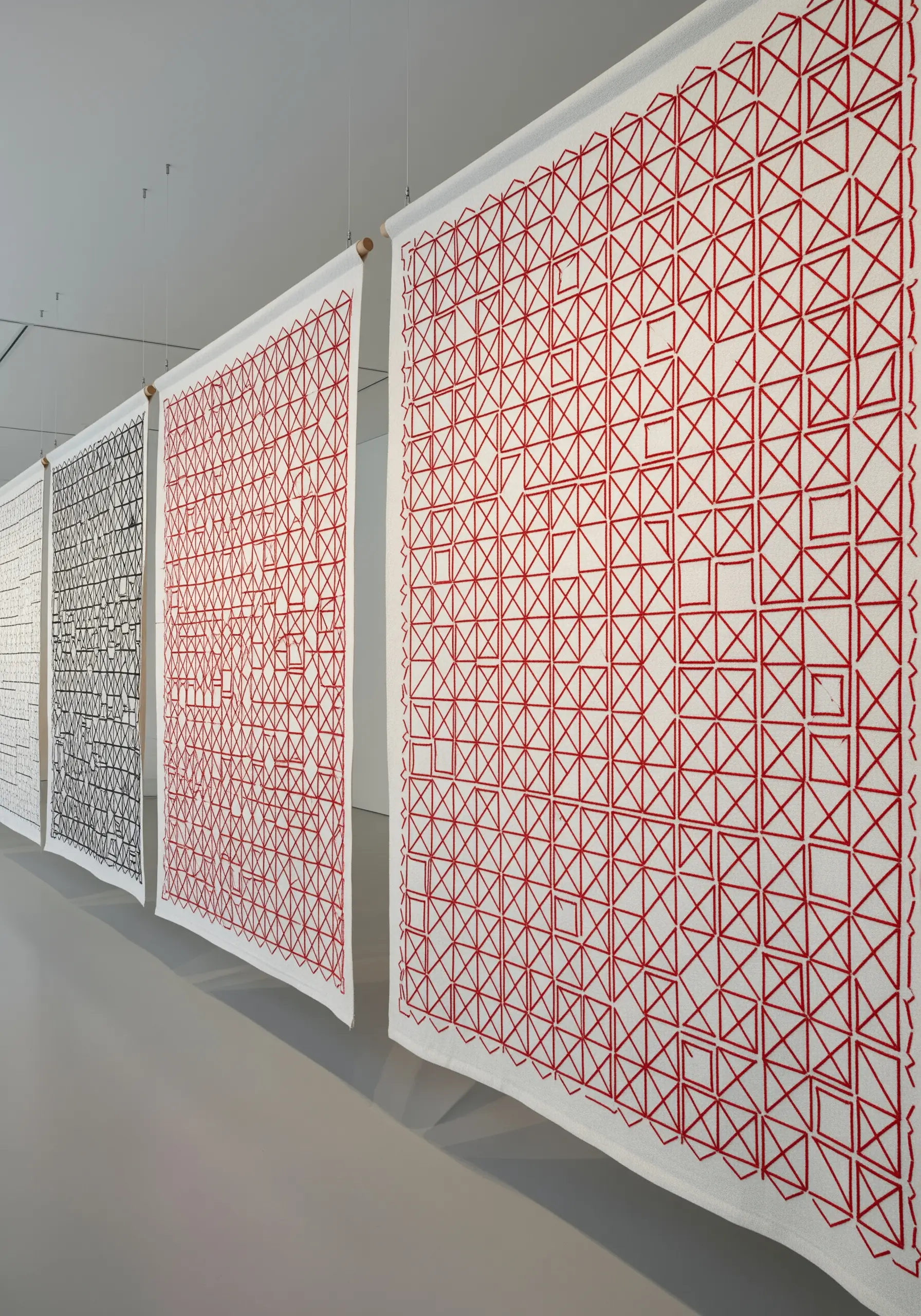 Large white fabric panels with repetitive red and black geometric line embroidery hanging in a gallery.