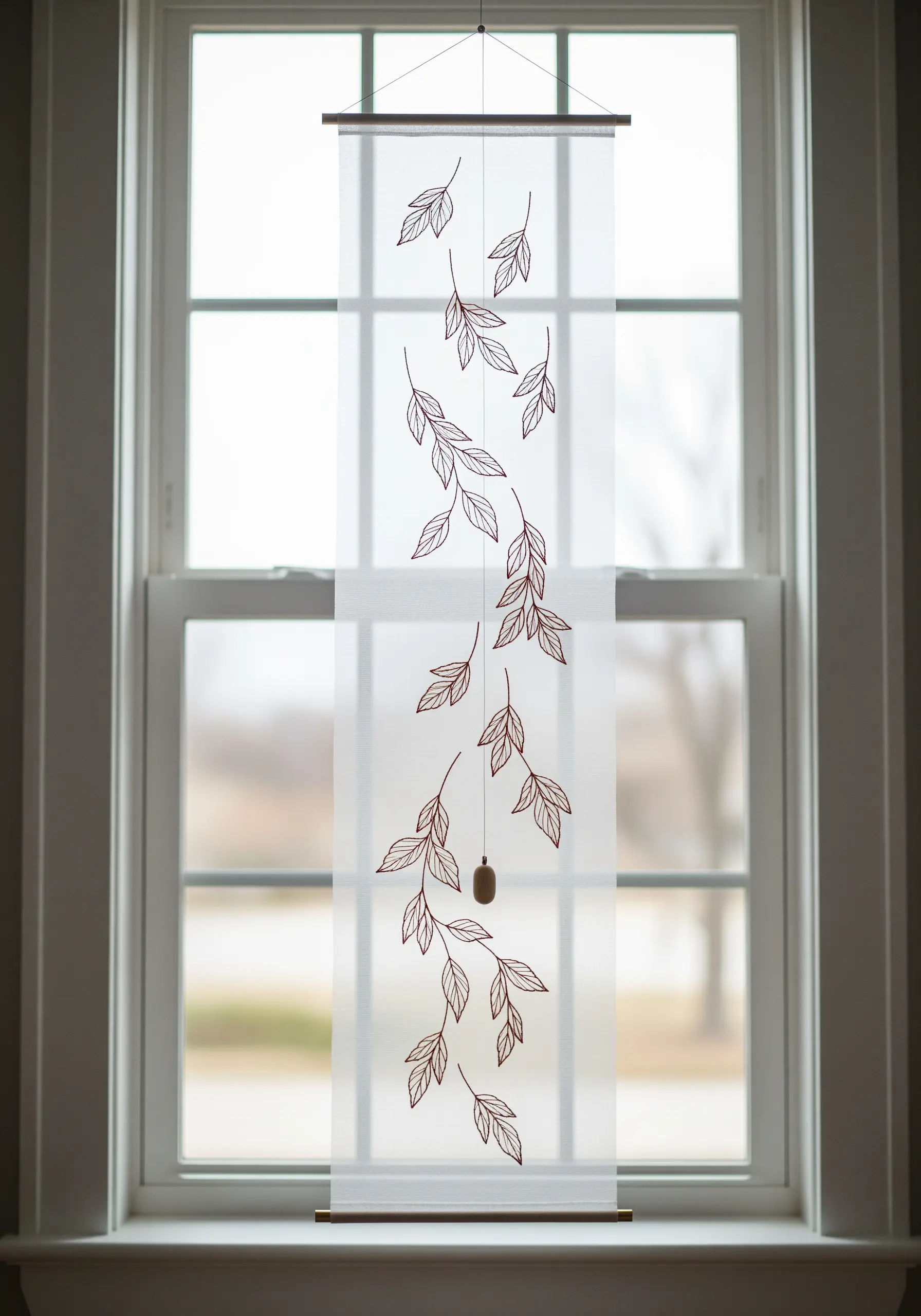 A sheer fabric wall hanging with embroidered outlines of cascading leaves, hanging in a window.