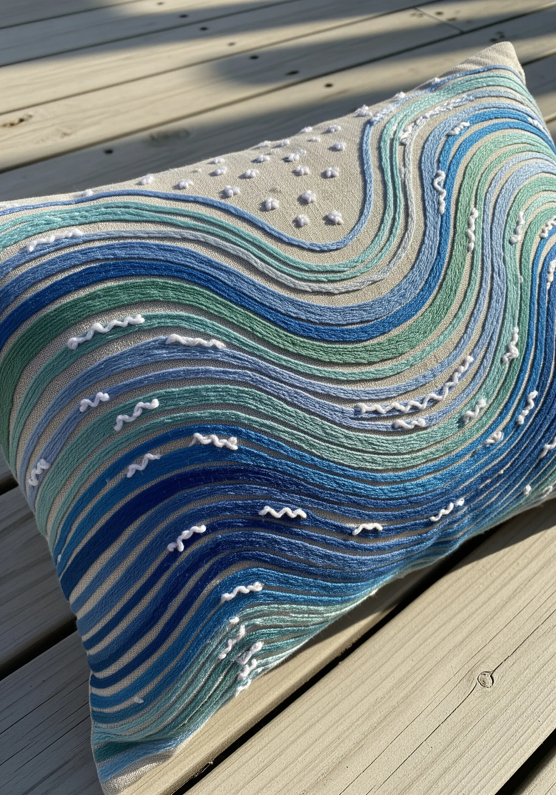 Abstract ocean waves embroidered on a pillow using couched blue and green yarn with white French knots.