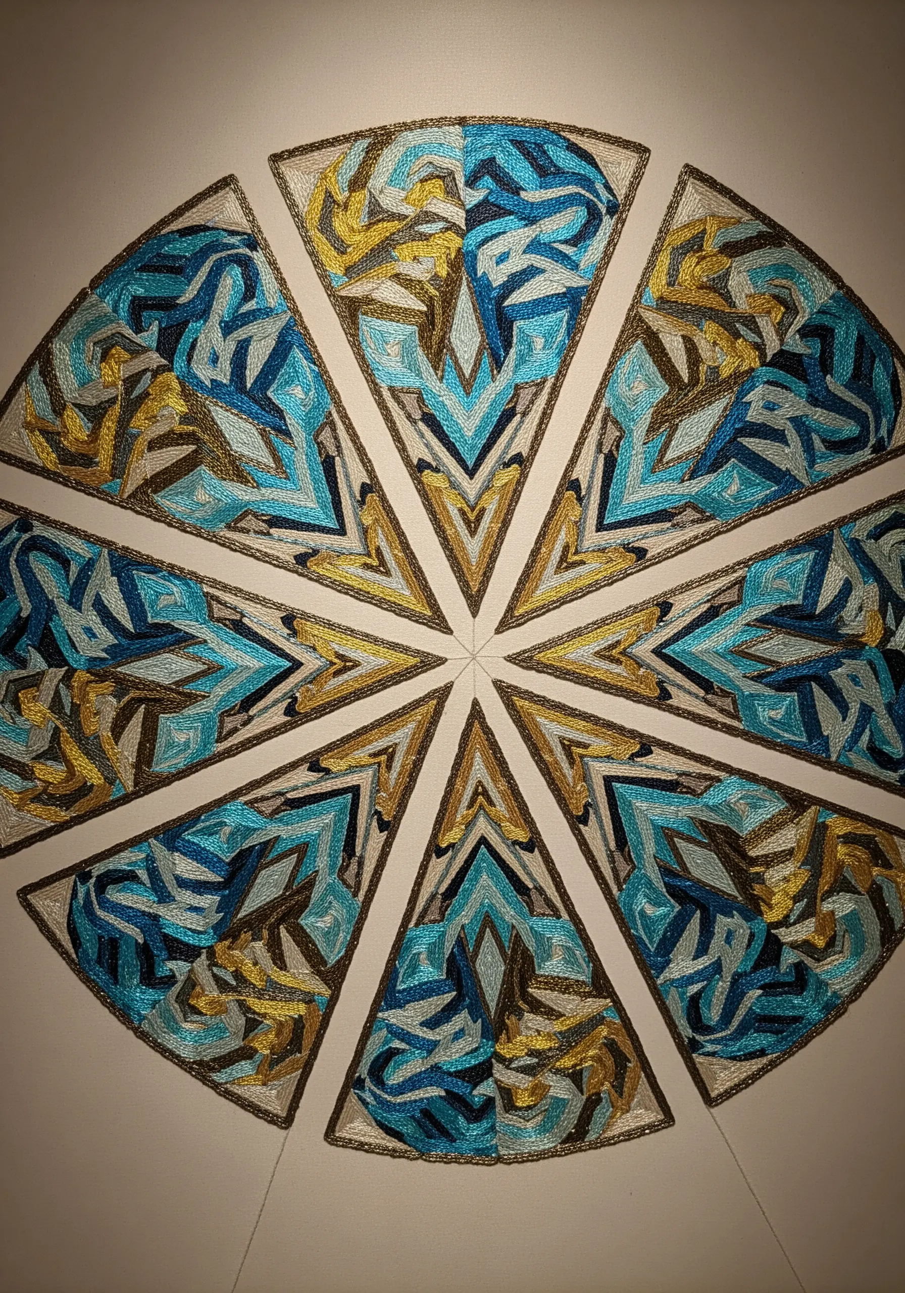A circular, kaleidoscopic design with geometric segments filled with abstract blue and gold stitches.