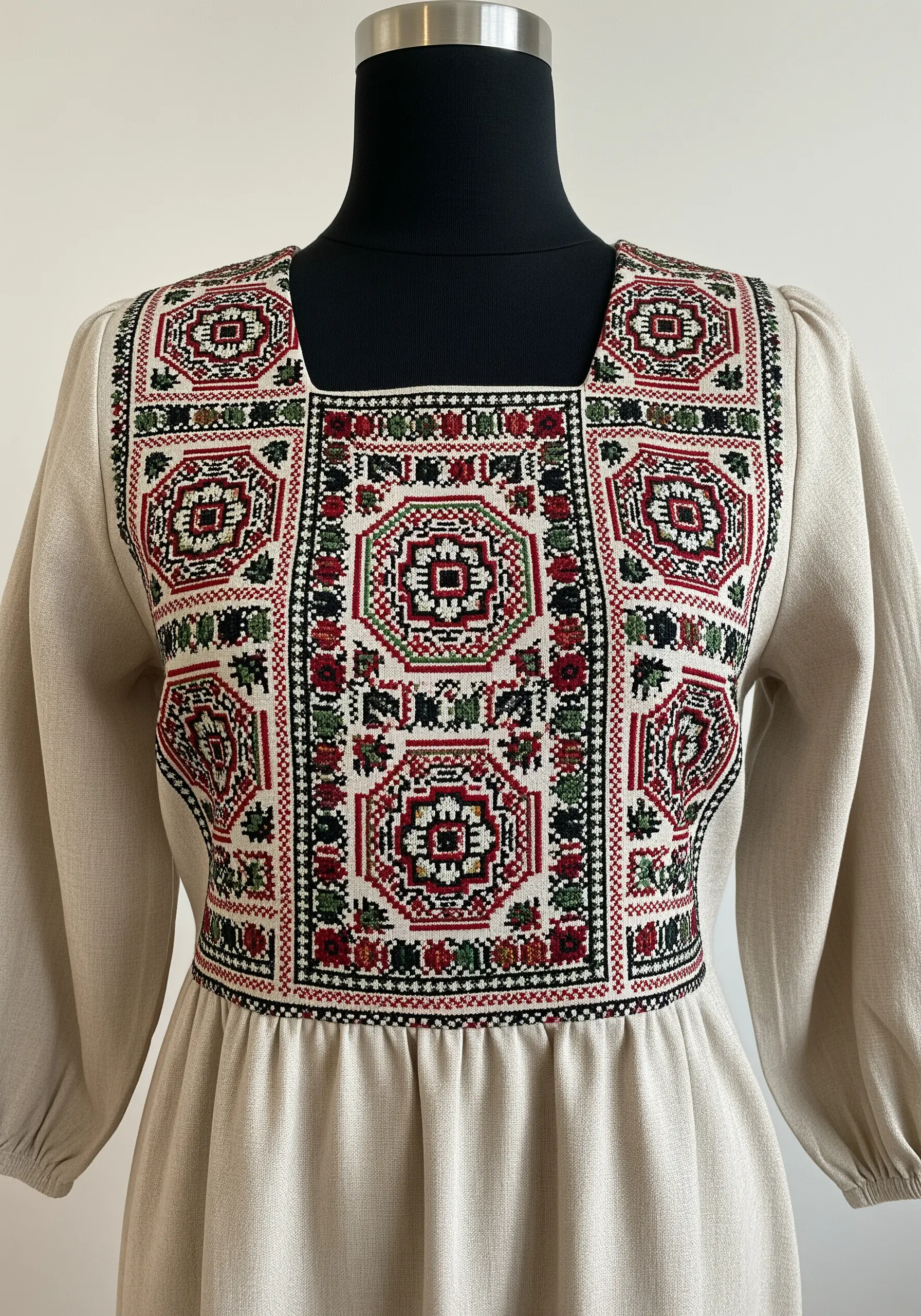 Geometric cross-stitch panel in red, green, and black on the bodice of a natural linen dress.