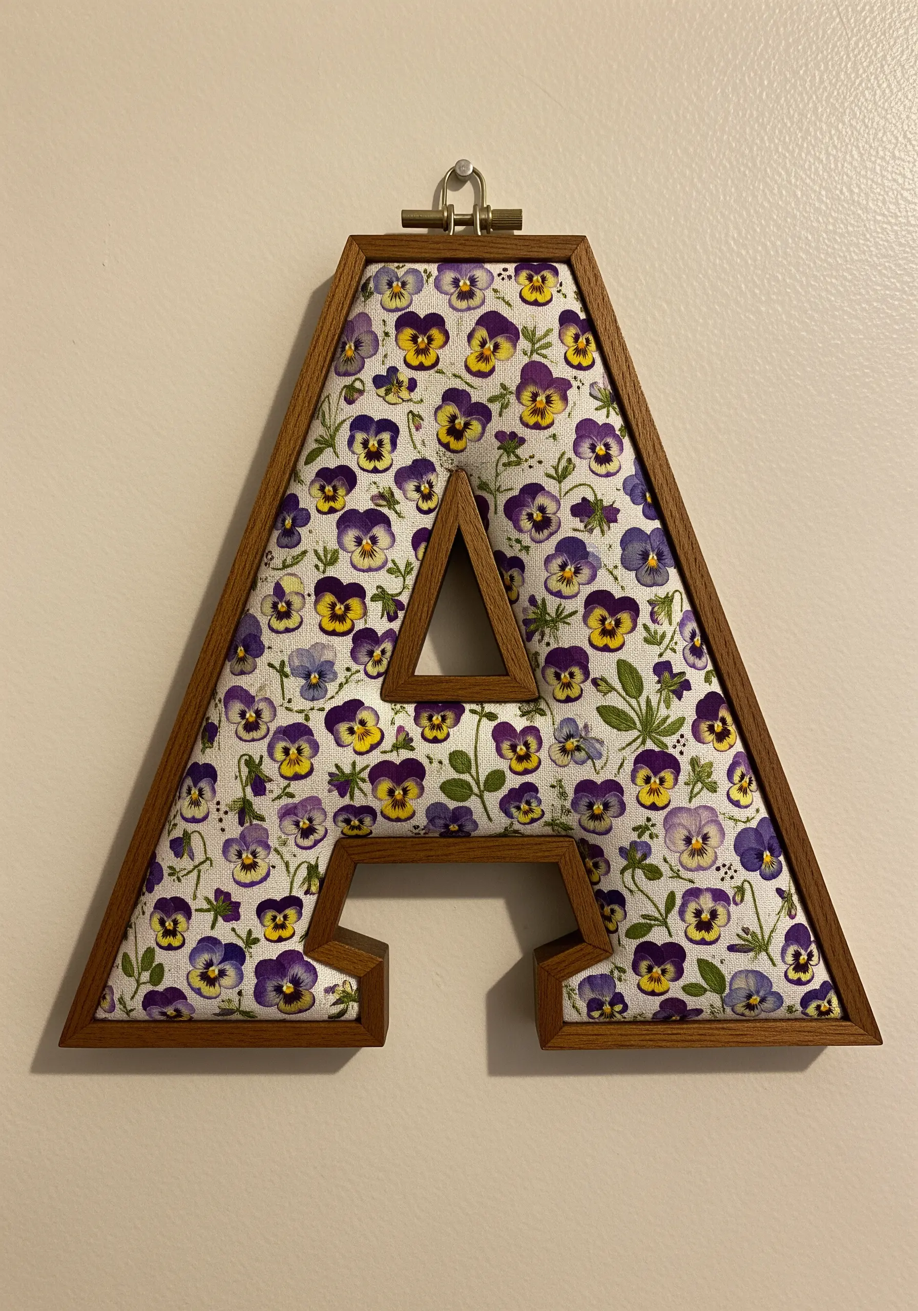 A wooden letter 'A' frame filled with a pansy-patterned fabric, hung on a wall.