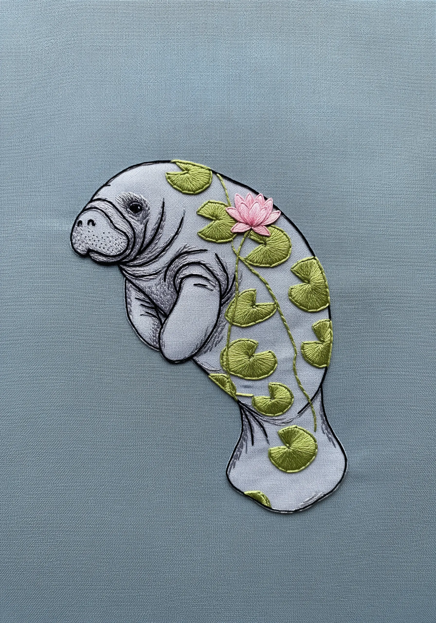 An embroidered manatee with delicate contour stitching, decorated with green lily pads and a pink flower.