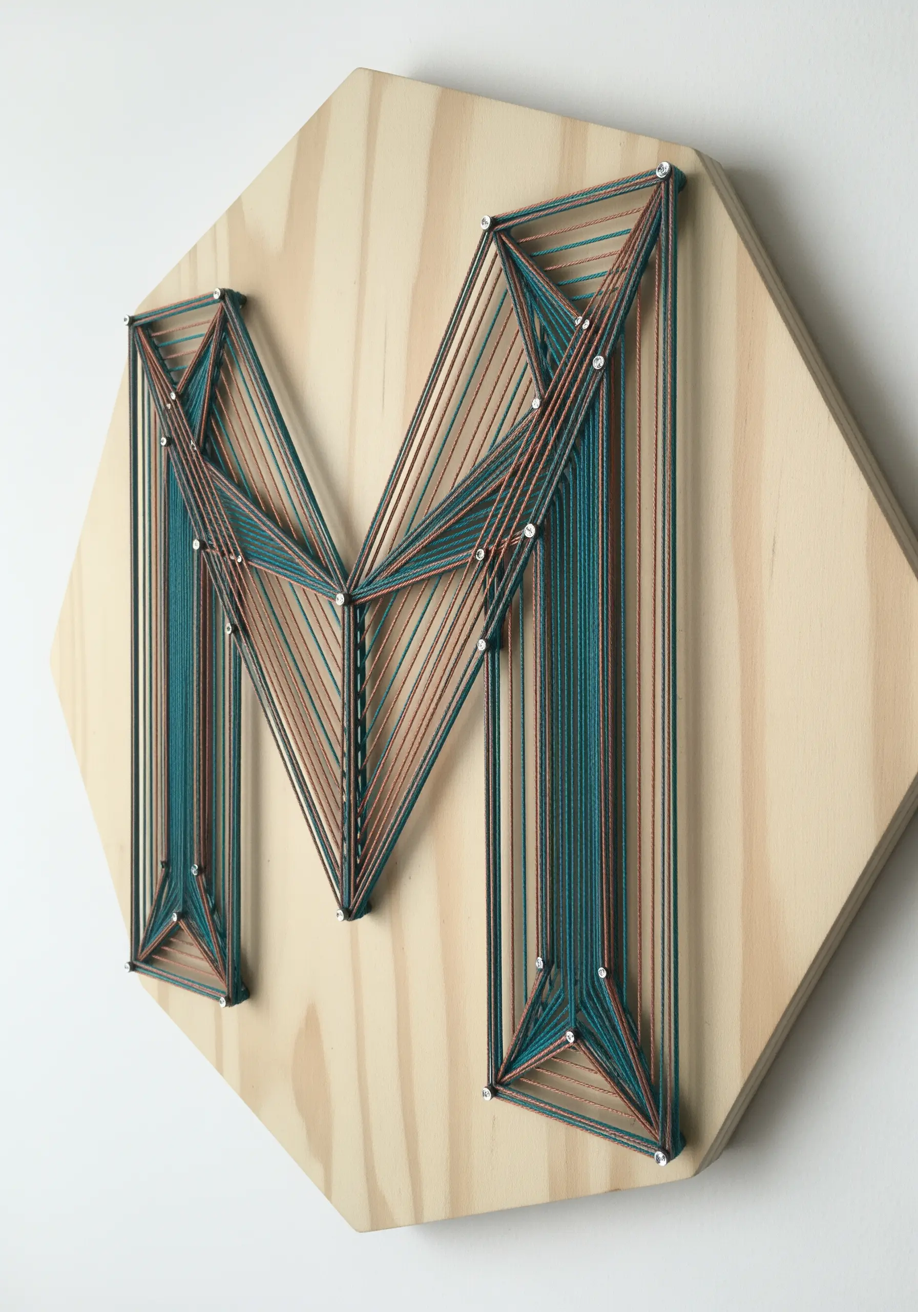 Teal and copper geometric string art of the letter M on a wood hexagon