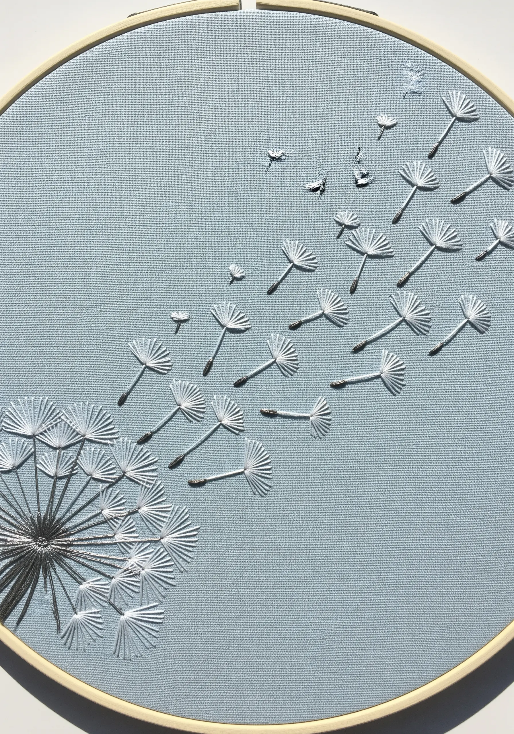Embroidery of a dandelion with seeds blowing away, stitched in white on a pale blue background.