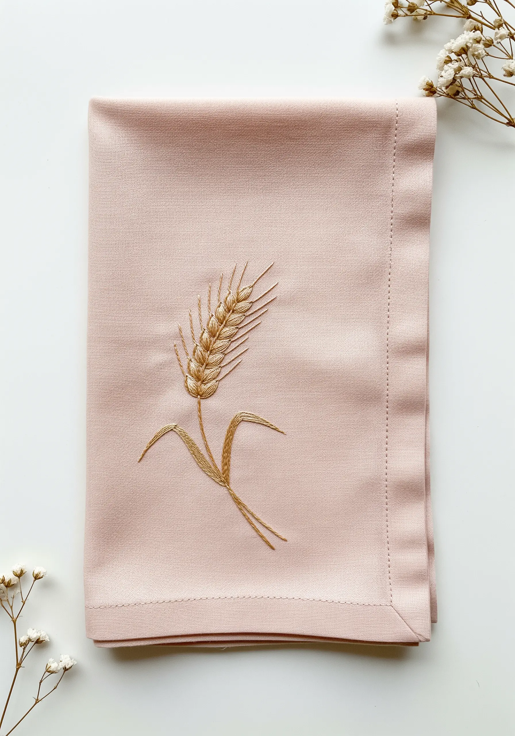A single wheat stalk embroidered in golden thread on a dusty pink napkin.