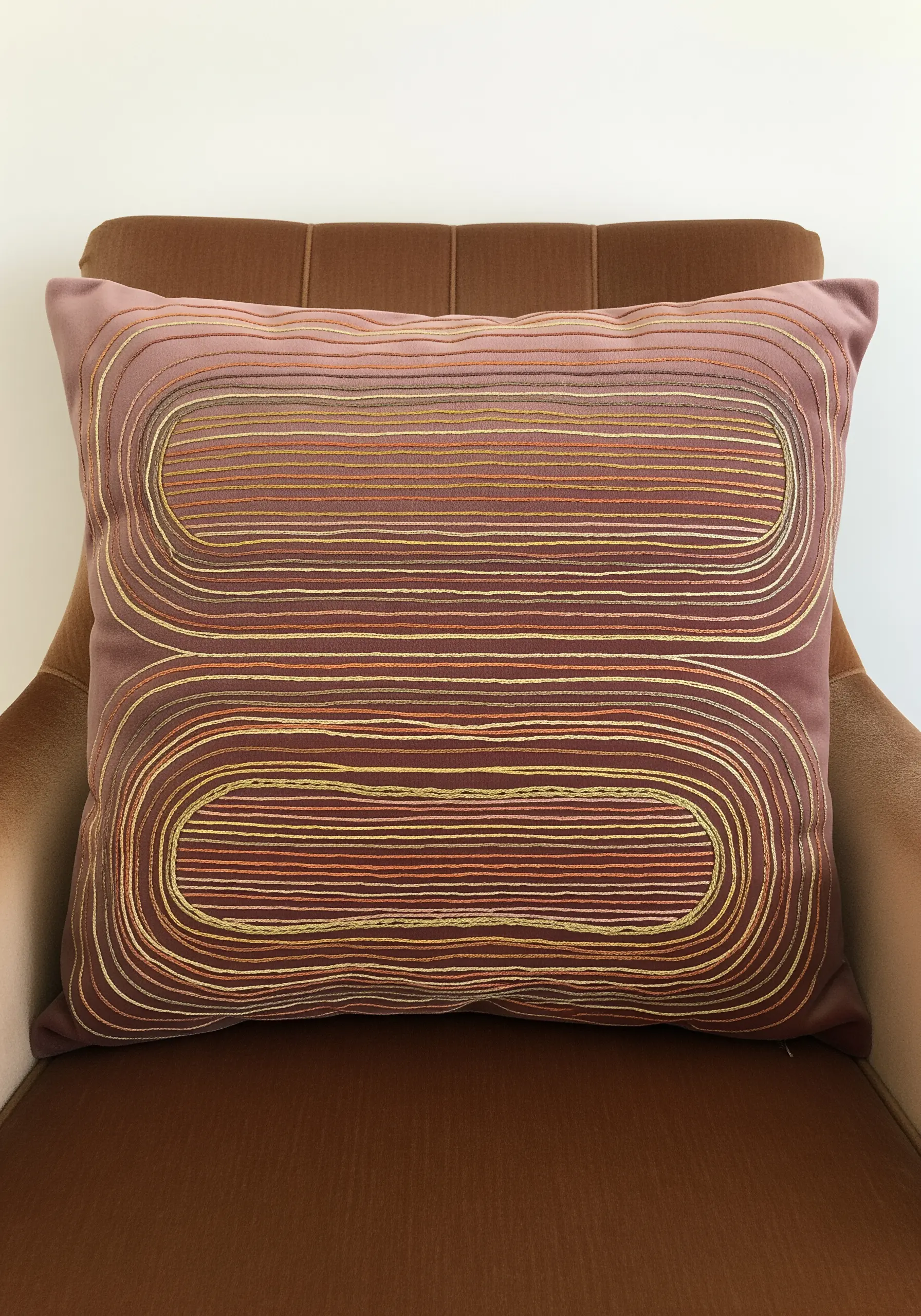 Concentric rounded rectangles in a warm, metallic thread gradient on a dusty rose pillow.